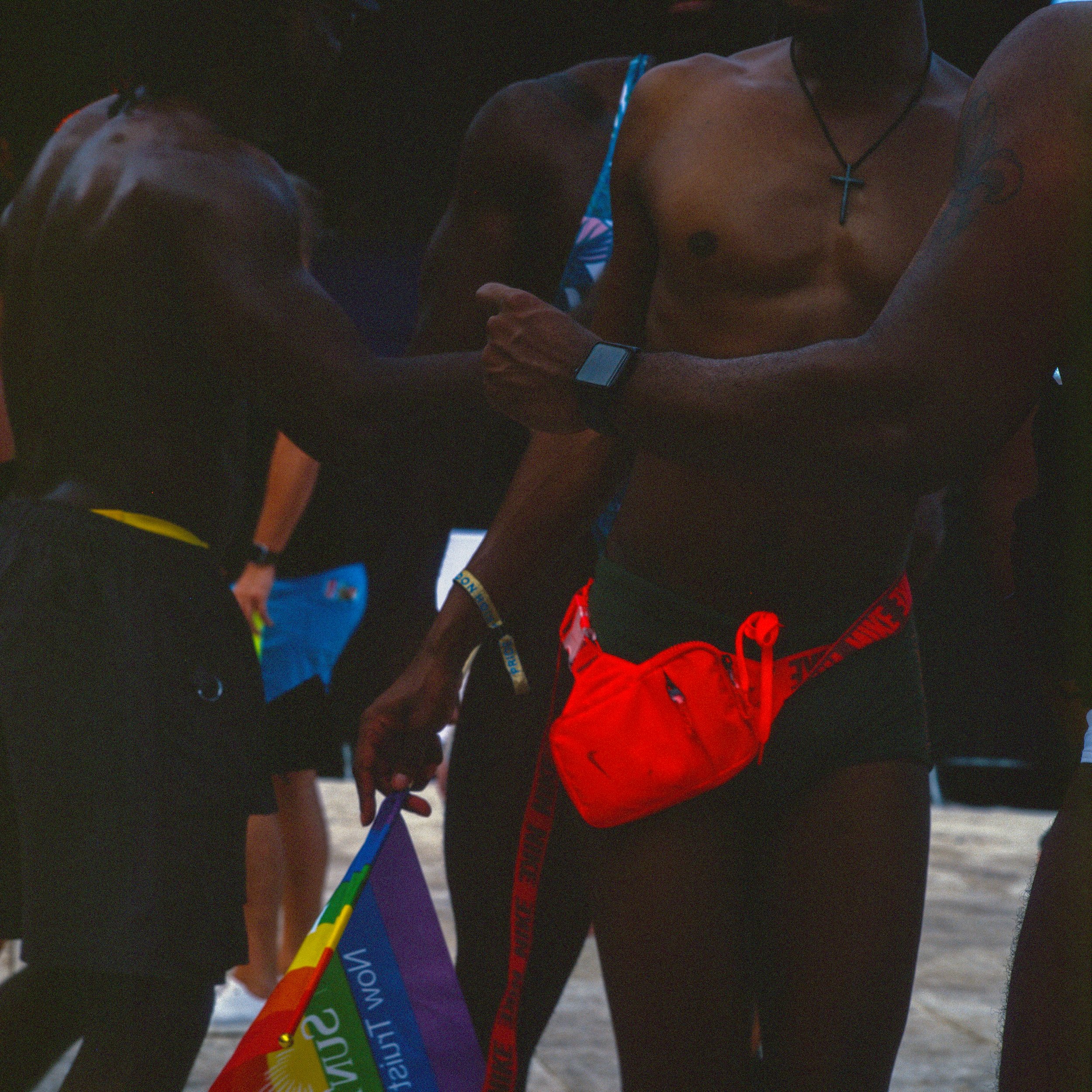 A shirtless man with a cross necklace and red waist bag, using a smartwatch, shaking hands with a person who is holding a rainbow pride flag at a beach event.