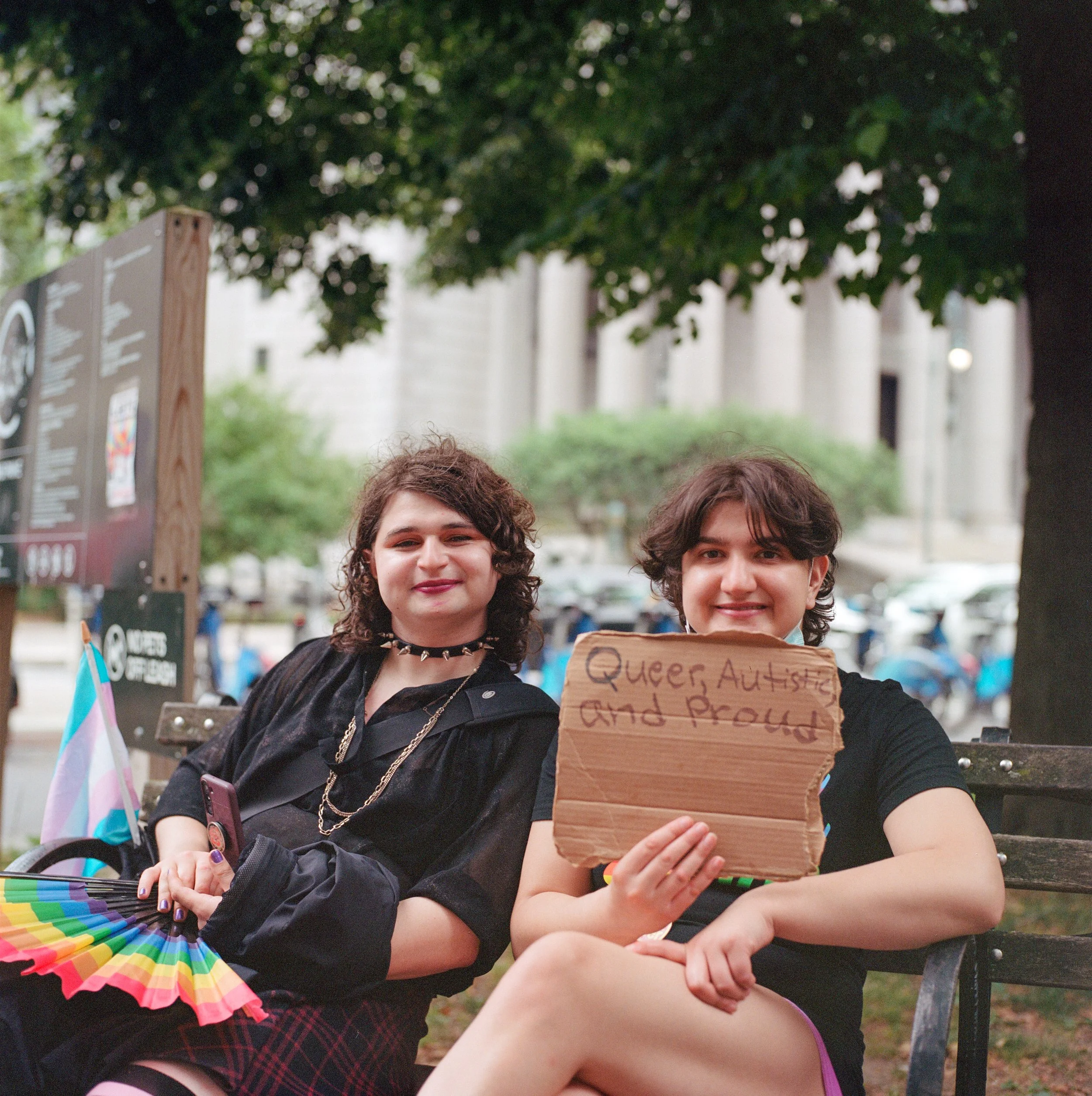 Two joyful LGBTQ+ individuals sitting on a park bench, holding a cardboard sign that reads "Queer, Autistic, and Proud." They are surrounded by trees and urban buildings, wearing dark clothing, with rainbow accessories and flags, during daytime.