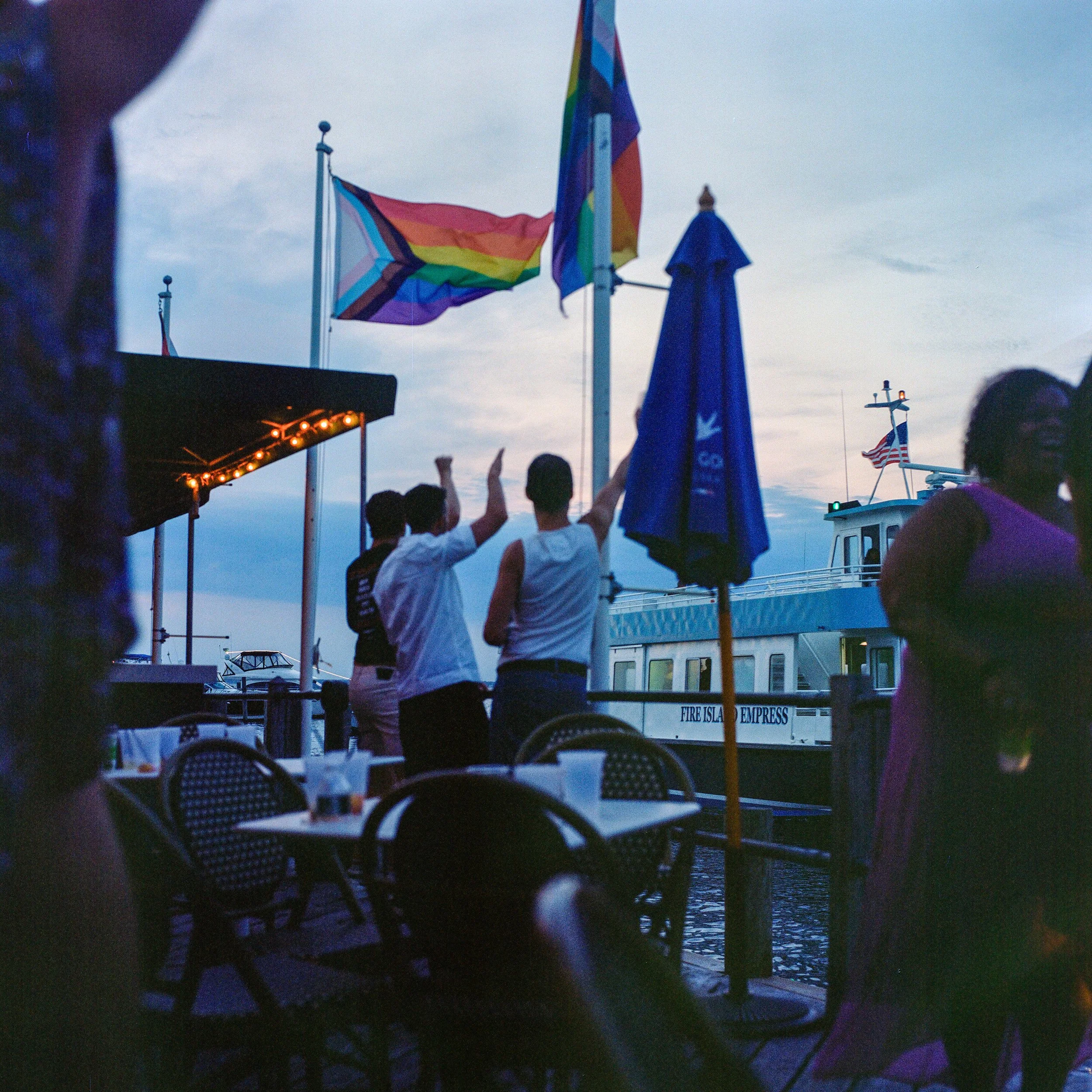 People dancing on a dock near a boat with a rainbow pride flag flying, during evening with cloudy sky.