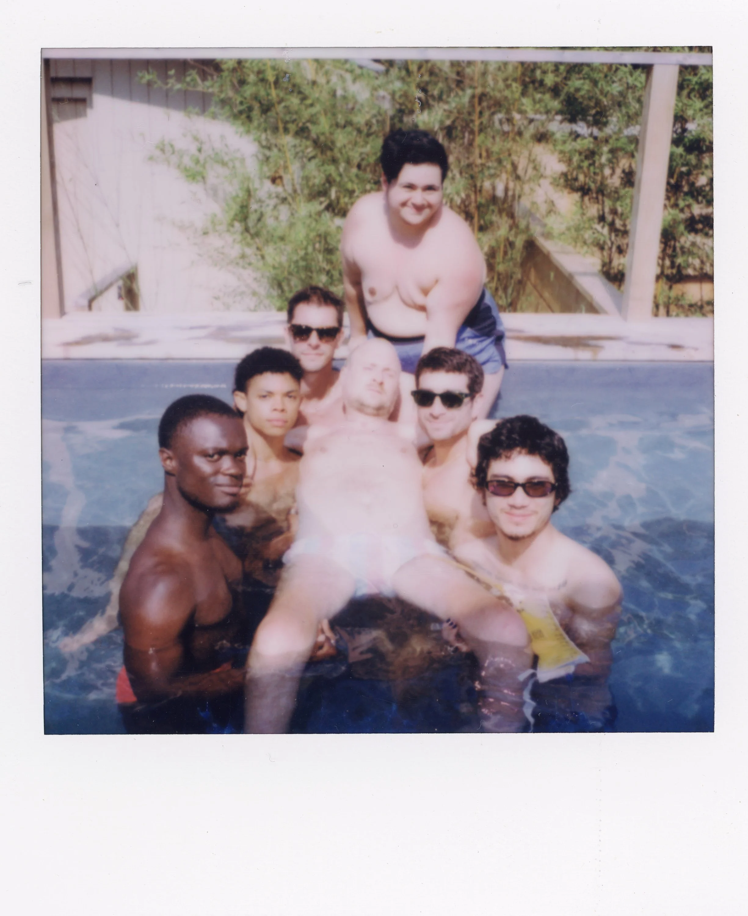 A group of seven men in a hot tub outdoors, some wearing sunglasses, with a wooden deck and greenery in the background.