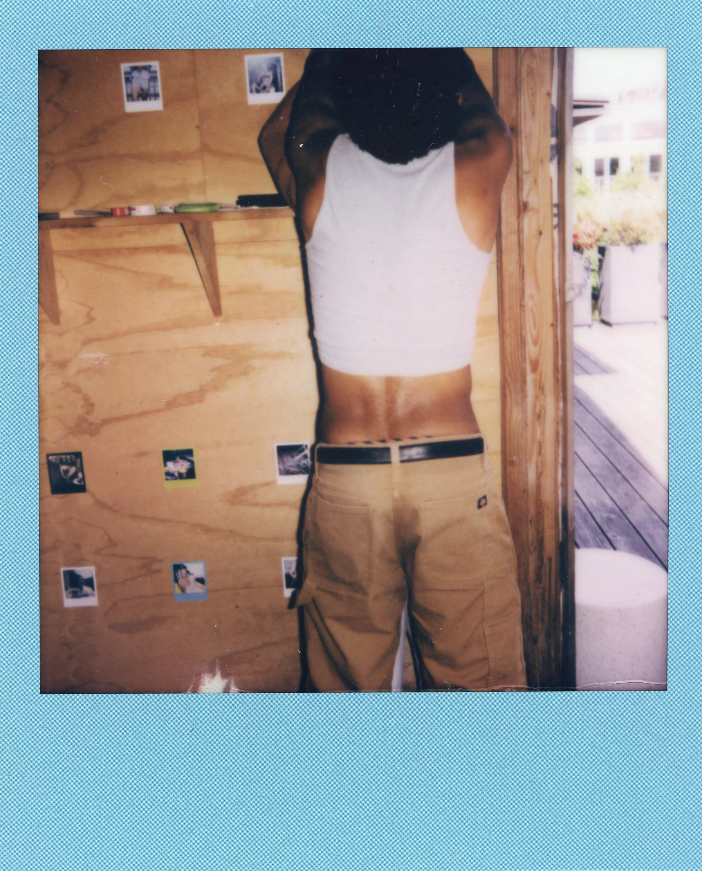 Photograph of a person seen in a mirror, with their face obscured, wearing a white tank top and khaki cargo shorts, standing against a wooden wall decorated with small Polaroid pictures.