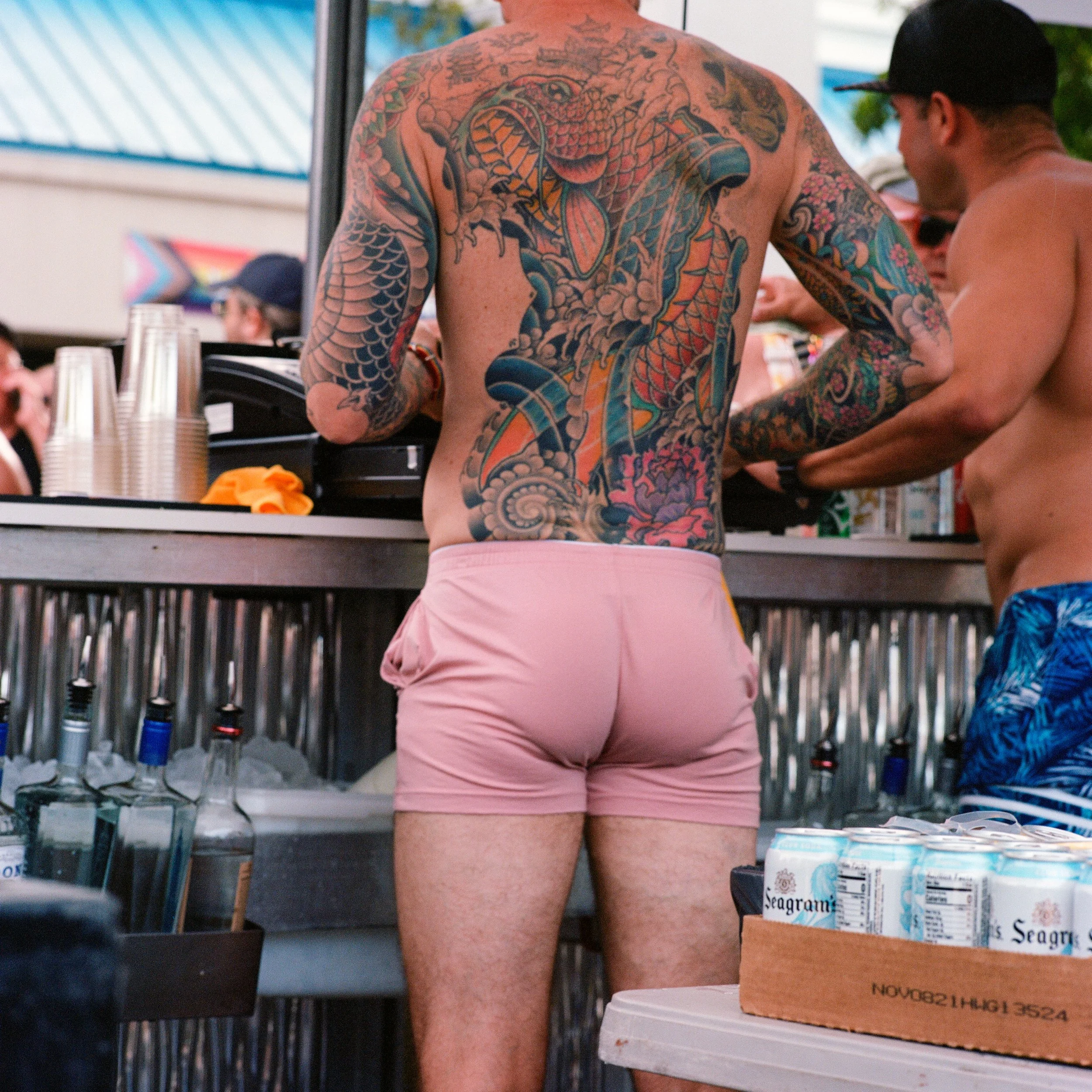 A shirtless man with heavily tattooed back and arms is at a bar, wearing pink shorts. People in the background are also shirtless, and there are bottles and cans on the bar and nearby table.