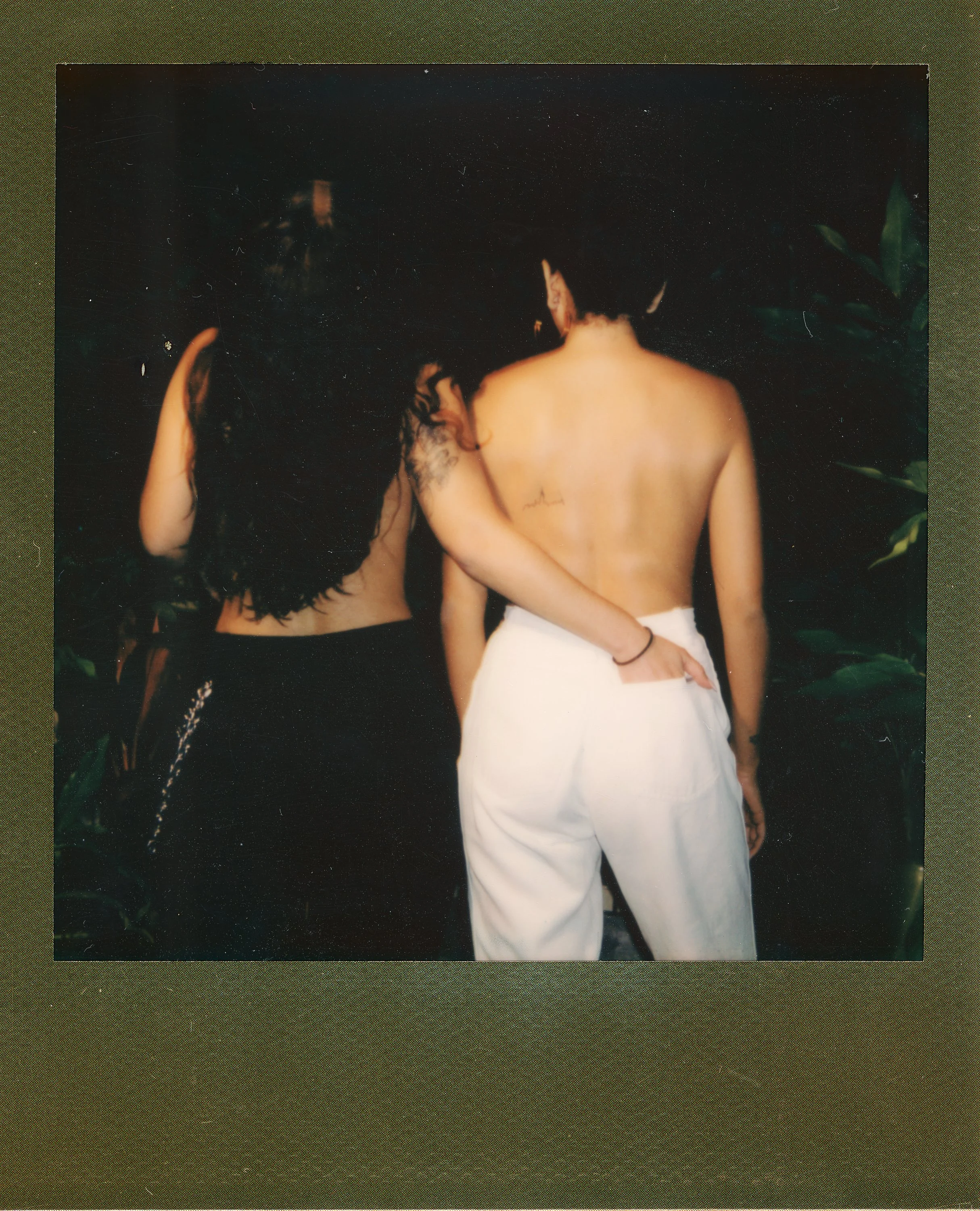Two people, one with long dark hair and the other with short dark hair and tattoos, standing side by side with their backs to the camera, surrounded by dark greenery.