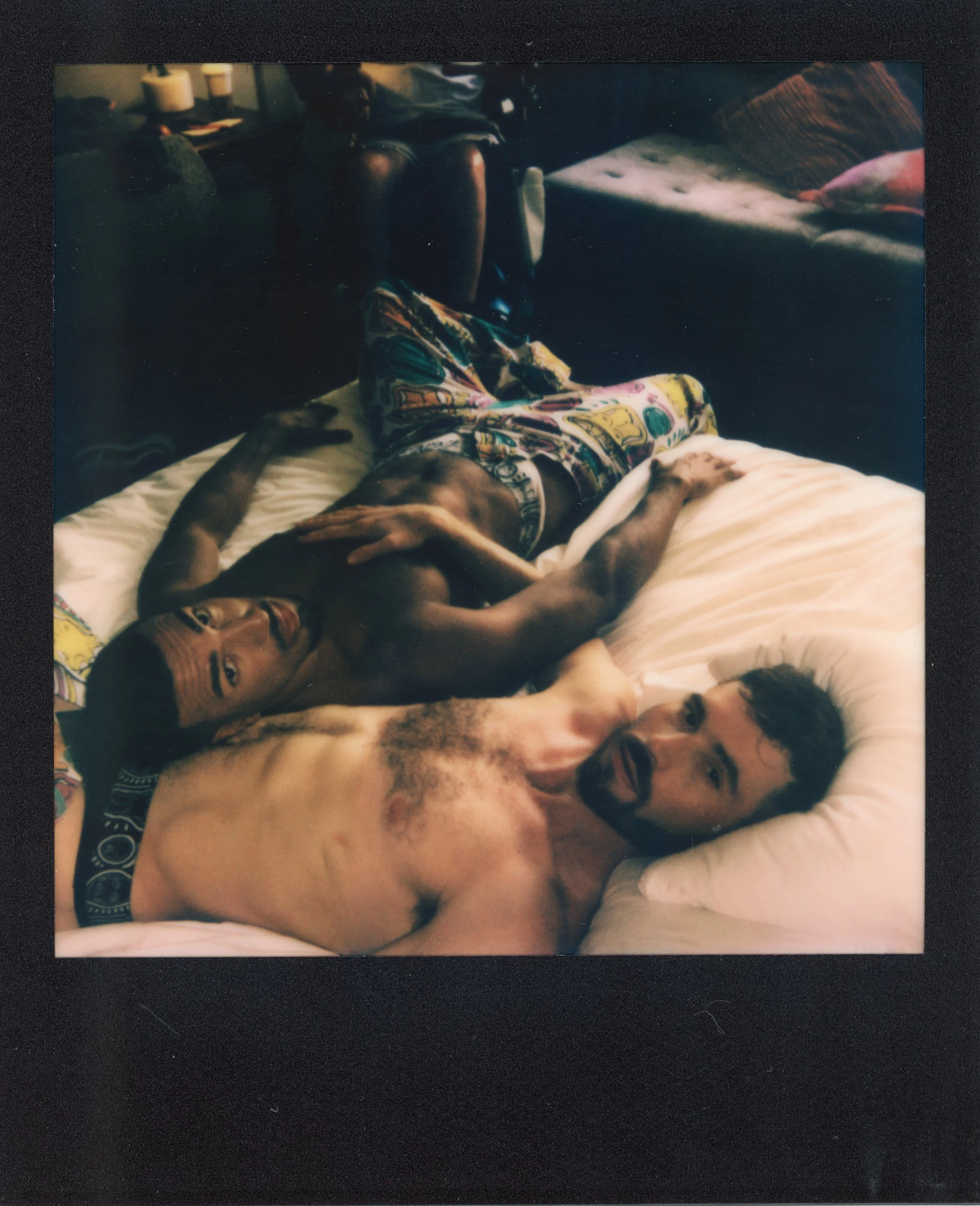 Two shirtless men lying on a bed with one looking at the camera and the other gazing upwards, surrounded by dim lighting and colorful furnishings.