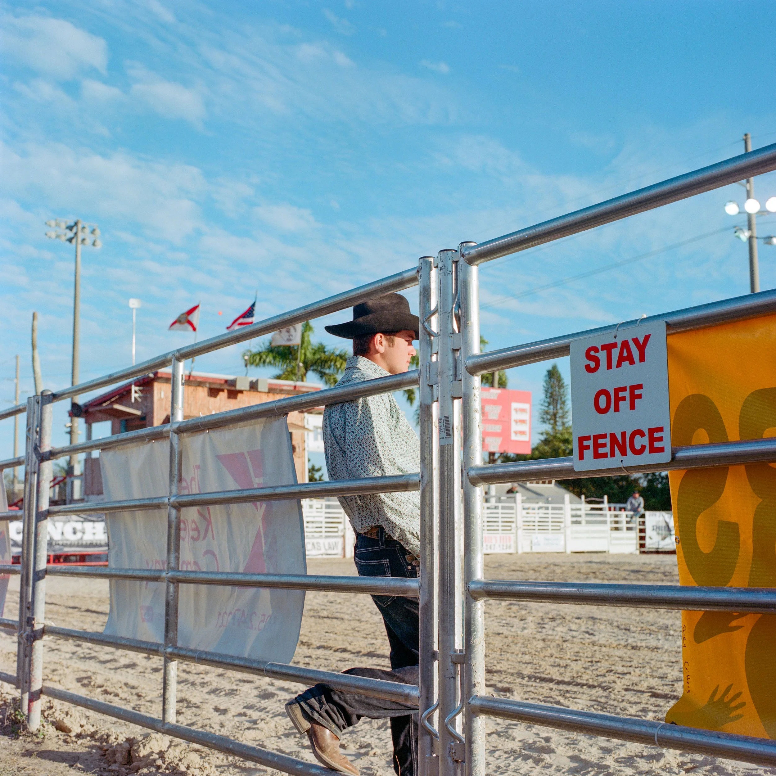 A man dressed as a cowboy standing behind a metal fence with a warning sign that reads 'Stay Off Fence' at a rodeo arena, under a partly cloudy sky.