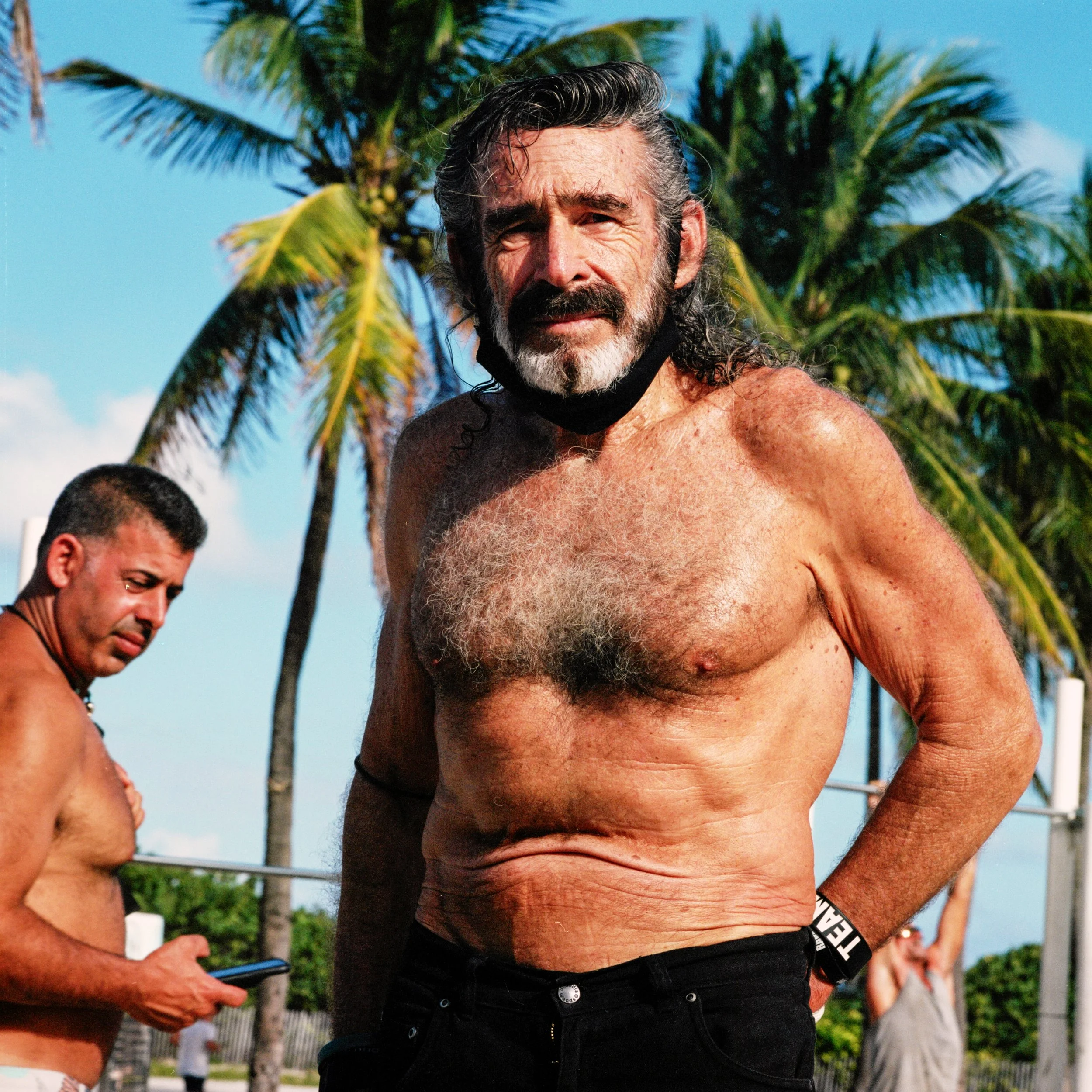 A shirtless middle-aged man with gray hair and beard standing outdoors in front of palm trees, with a blue sky background, looking at the camera.