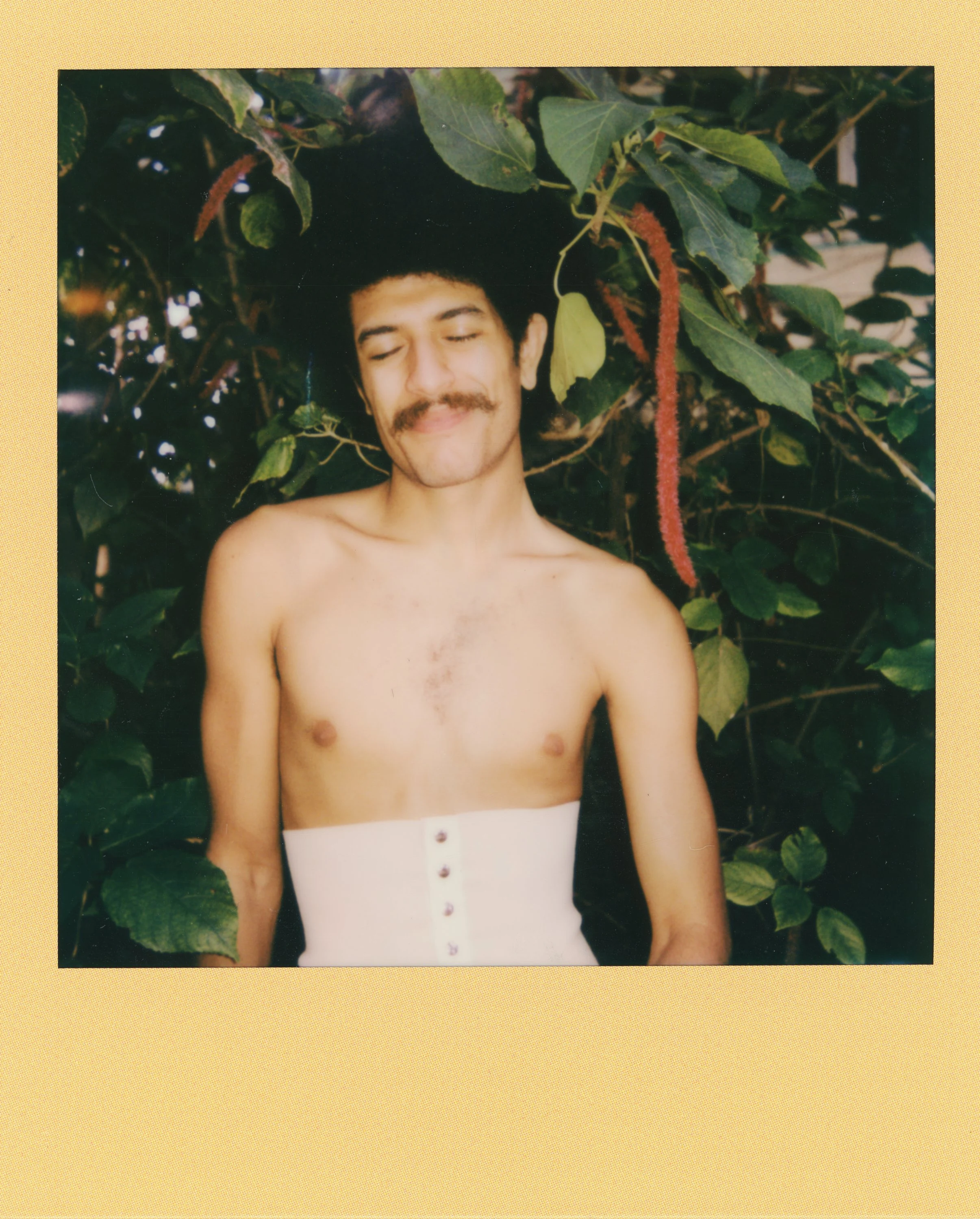 A shirtless man with a mustache and closed eyes, standing in front of leafy green plants with red flowers, smiling gently.