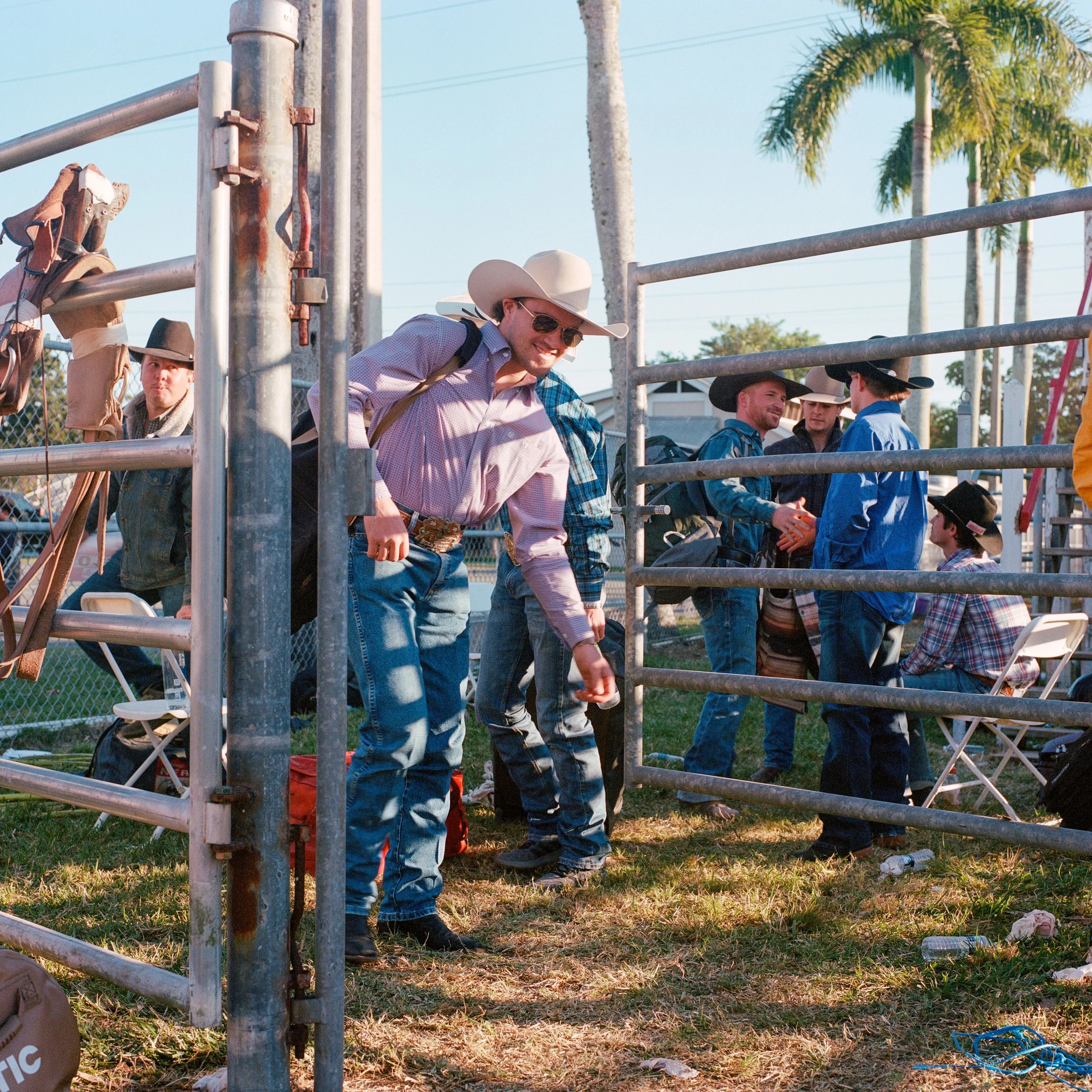 Group of men dressed in western attire, including cowboy hats, gathered outdoors near metal fencing, engaging in conversation and greeting each other on a sunny day with palm trees in the background.