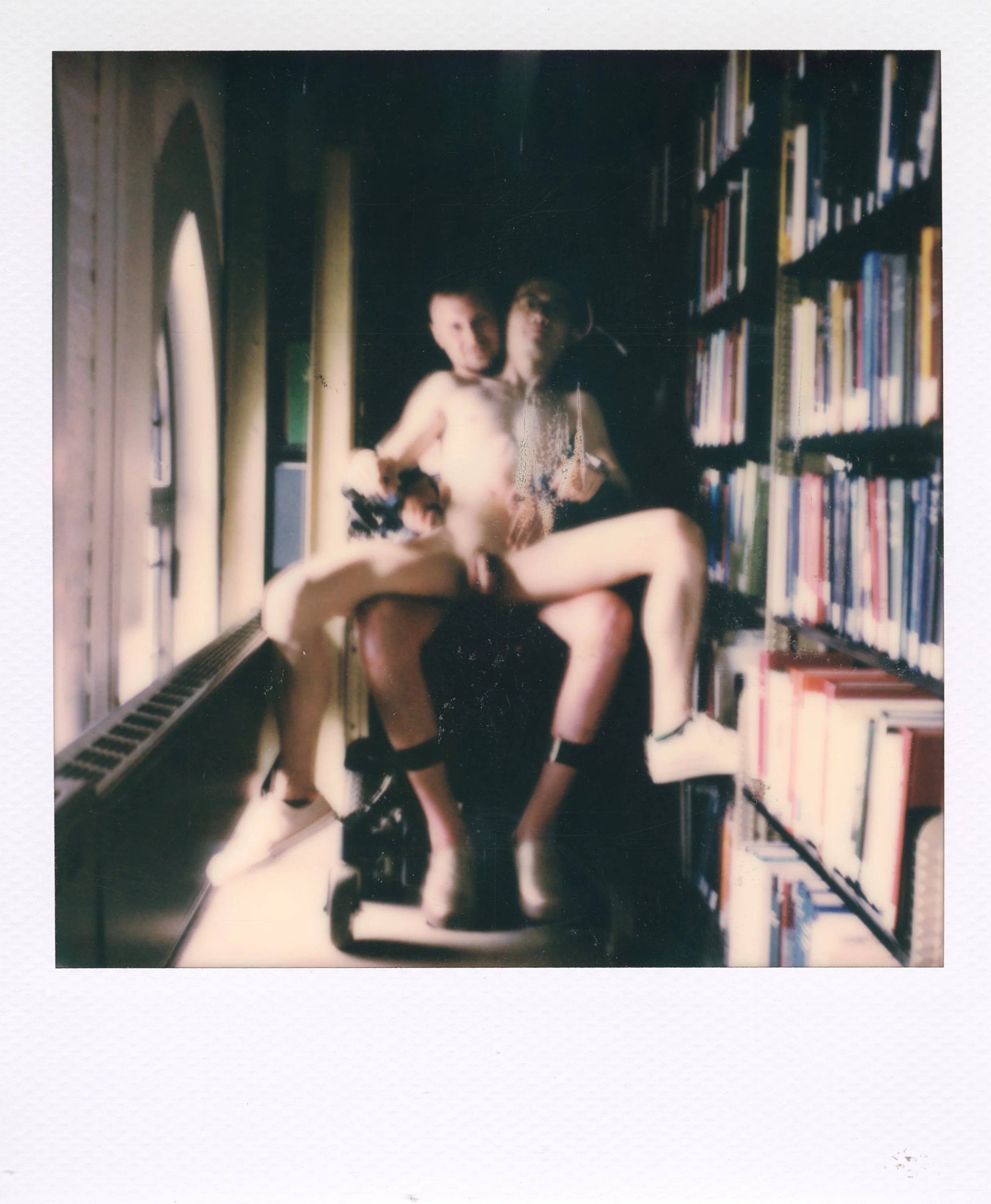 Two people sitting on a bookshelf in a room with large windows, with one person sitting on the other's lap. Both are partially nude and wearing socks, with one wearing boots. The room appears to be a library or bookstore.