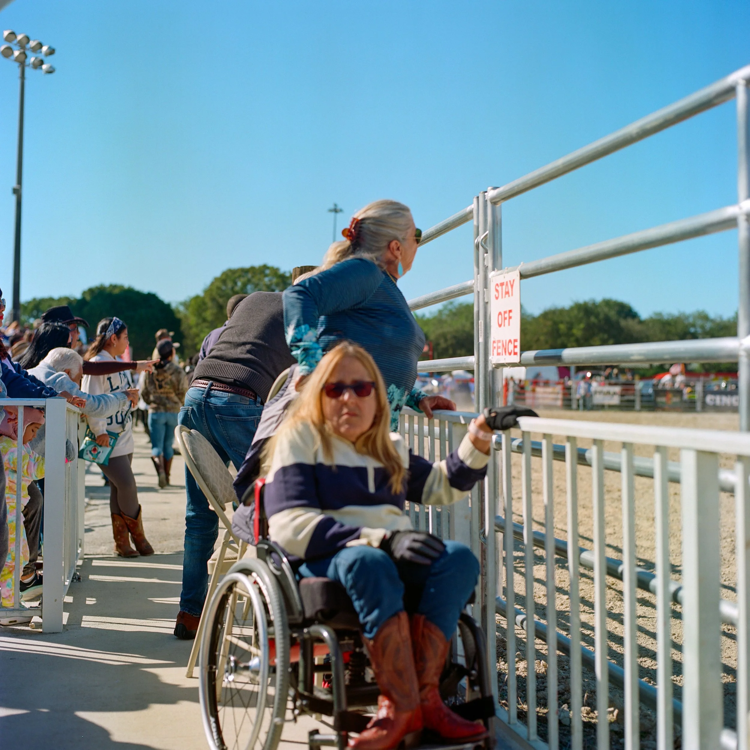 Crowd of people at an outdoor event, some leaning on a barrier, watching an activity on the field, with clear blue sky and trees in the background.