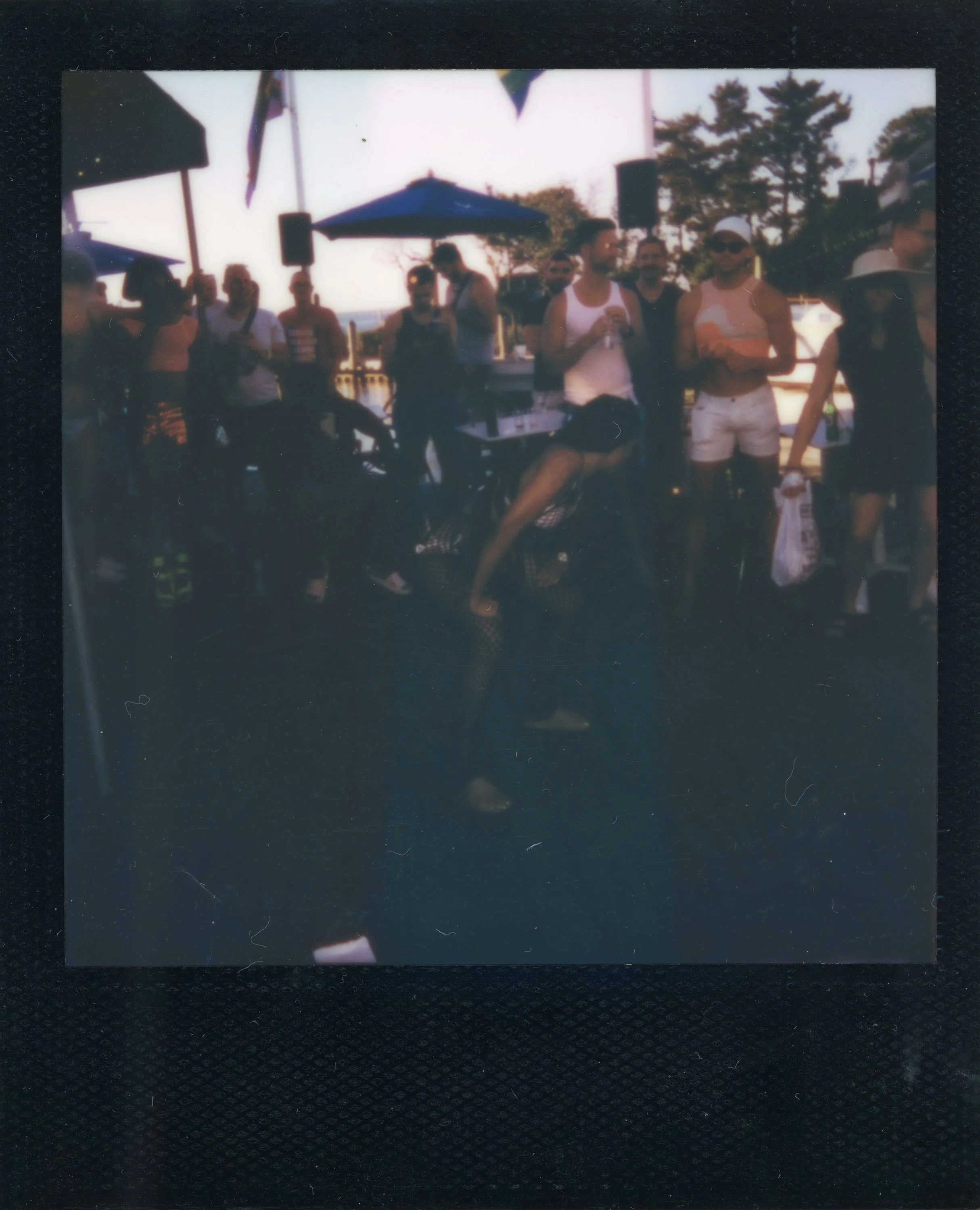 A group of people at an outdoor gathering under umbrellas, with some wearing casual summer attire, in a setting with trees in the background.