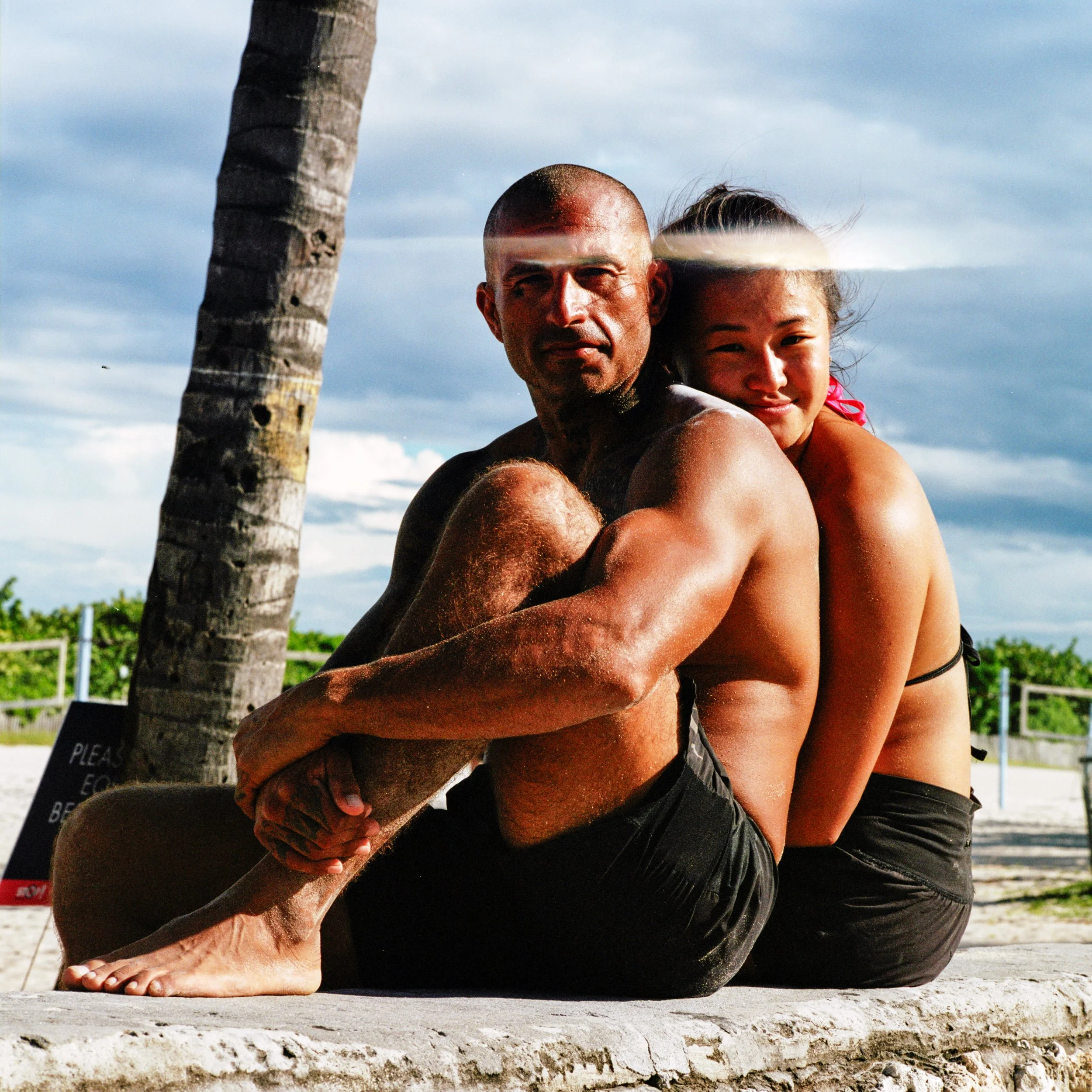 A man and a woman sitting on a low wall on a beach, with a palm tree nearby, in sunny weather.