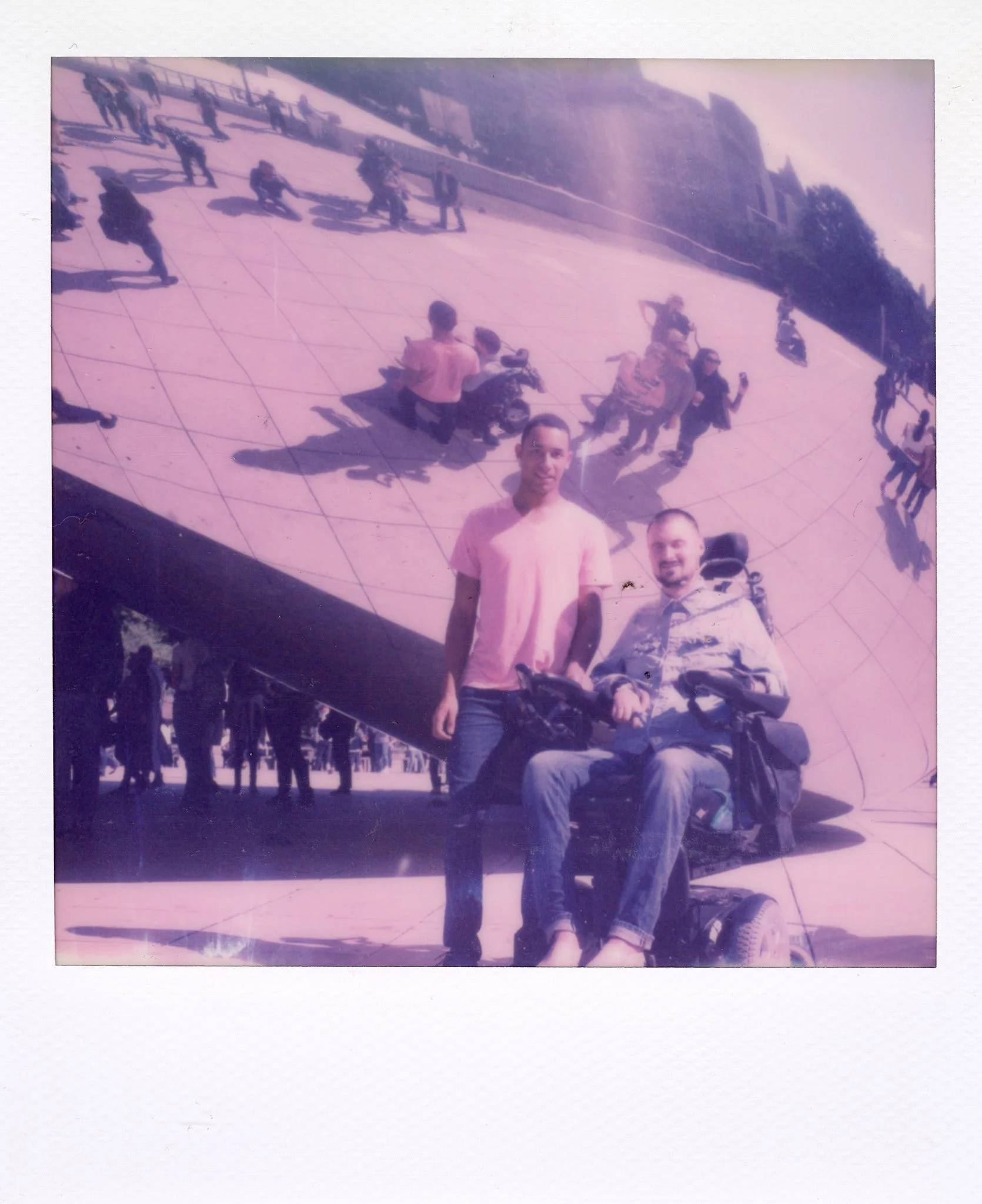 Two young men, one standing and one seated in a motorized wheelchair, pose in front of a mirrored structure with other people walking and riding bikes in the background.