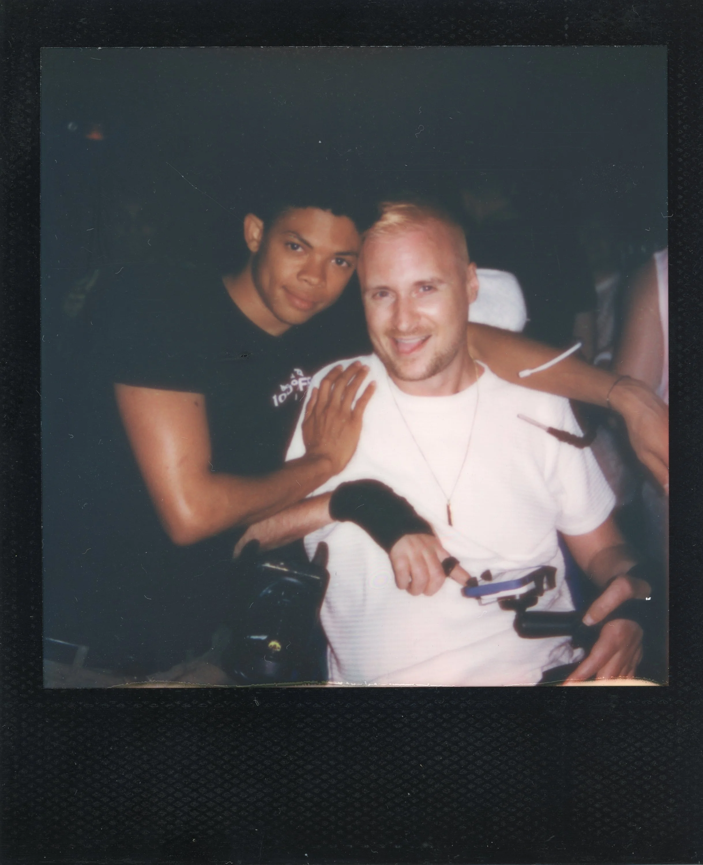 Two young men, one with dark skin and curly hair and the other with light skin and short blond hair, smiling and posing together. The man with light skin is seated in a wheelchair, wearing a white T-shirt, and the other man is standing beside him wit
