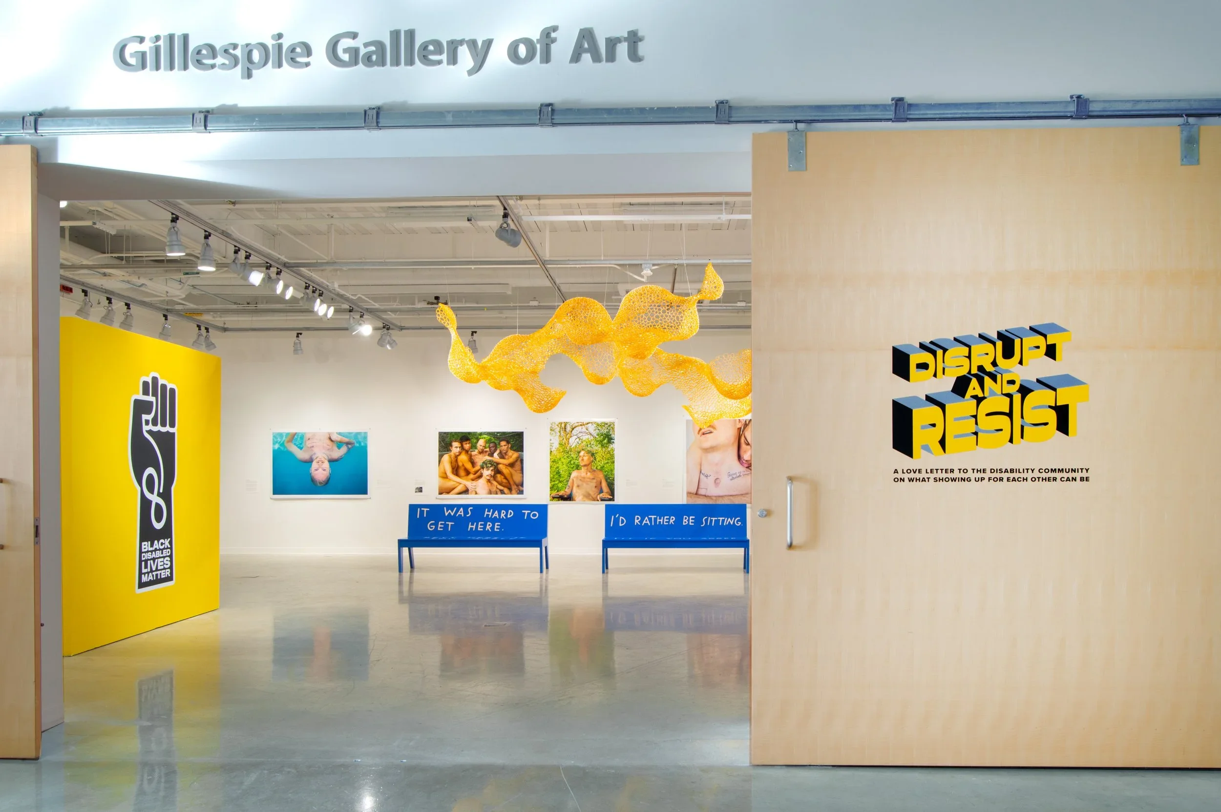 Entrance to Gilespie Gallery of Art with a yellow sign on the left displaying a raised fist and '#BlackDisabledLivesMatter' symbol. Inside the gallery, there are photographs, two blue benches with signs reading 'It was hard to get here' and 'I'd rather be sitting,' and a yellow sculptural installation hanging from the ceiling. On the right wall, there is a sign that says 'Disrupt and Resist' with a subtitle about a love letter to the disability community.