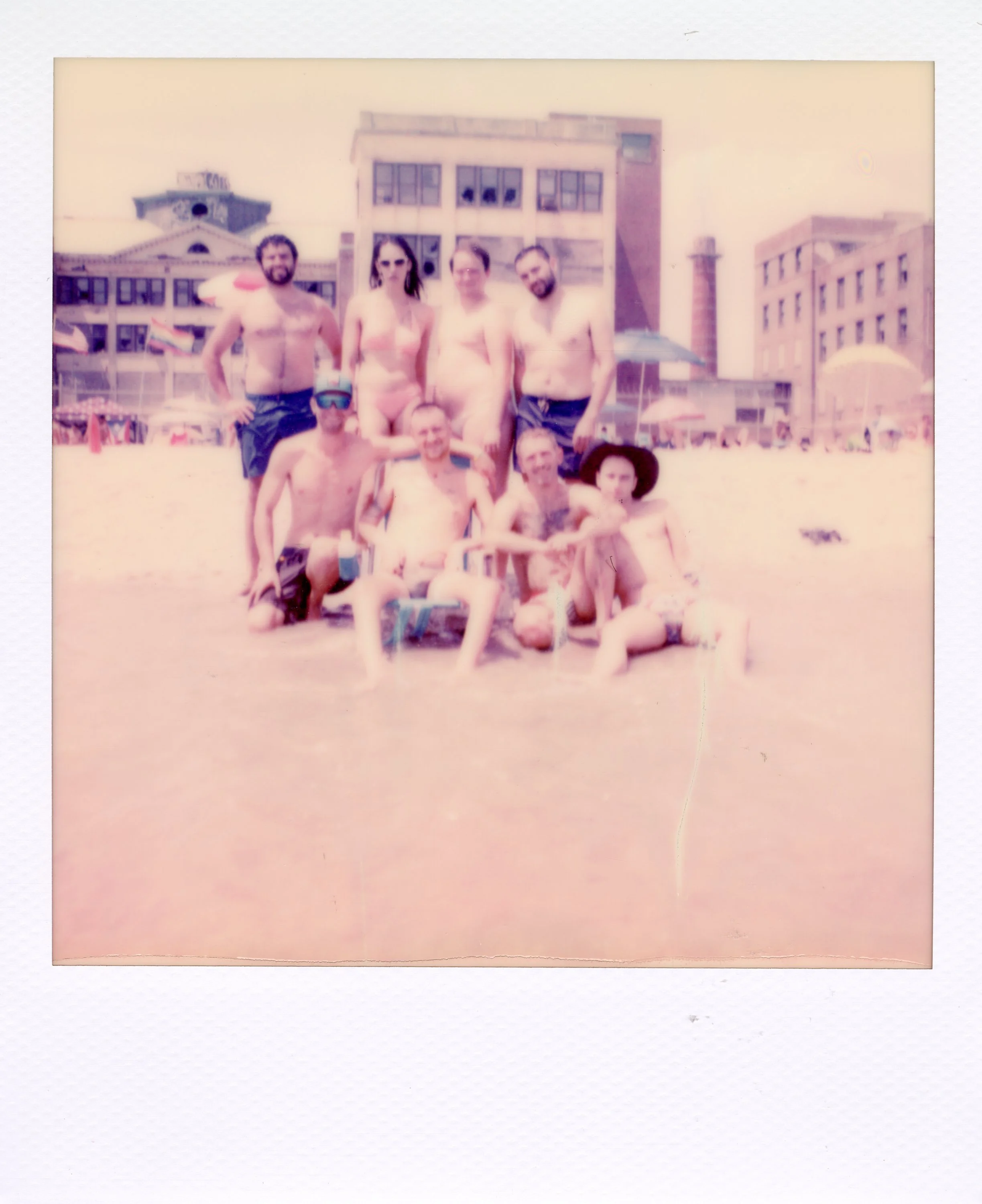 A group of people in swimsuits at the beach, some sitting in the water and others standing behind them, with buildings and beach umbrellas in the background.