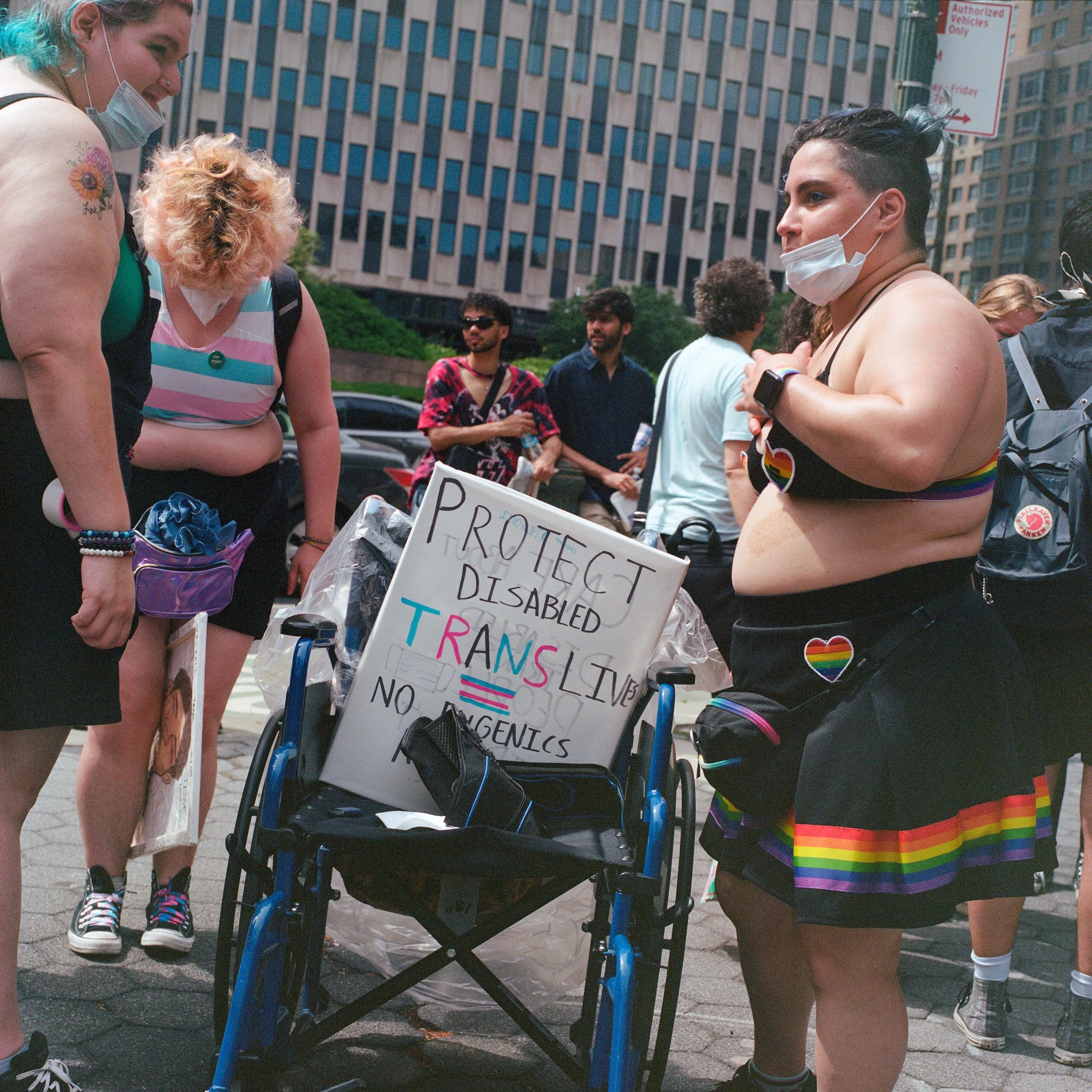 A group of people participating in a pride event, with one person in a wheelchair holding a sign that reads 'Protect Disabled Trans Live No Eugenics'. The individuals are dressed in rainbow-themed clothing and accessories, and some are wearing face m