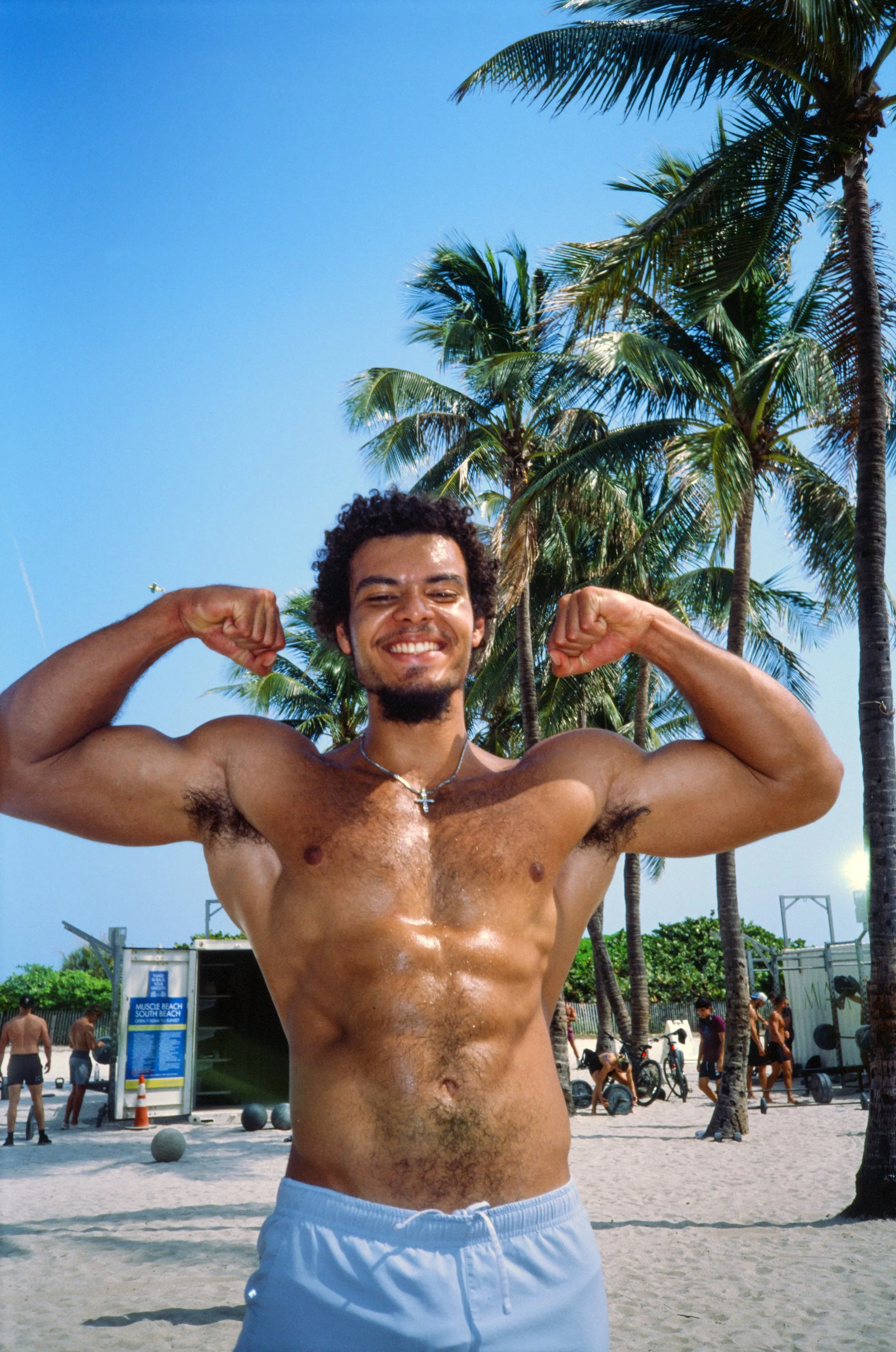 A young man with curly hair and a beard, wearing a cross necklace, smiling and flexing his biceps at the beach, surrounded by palm trees and other people in the background.