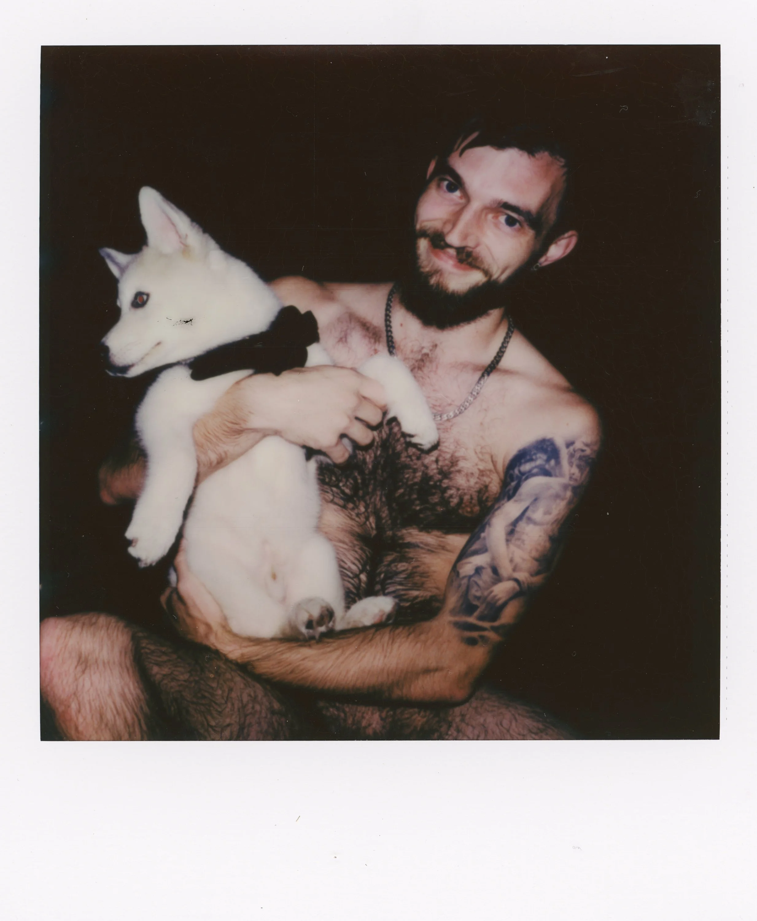 A shirtless man with tattoos holds a white and brown puppy with a black collar against a dark background.
