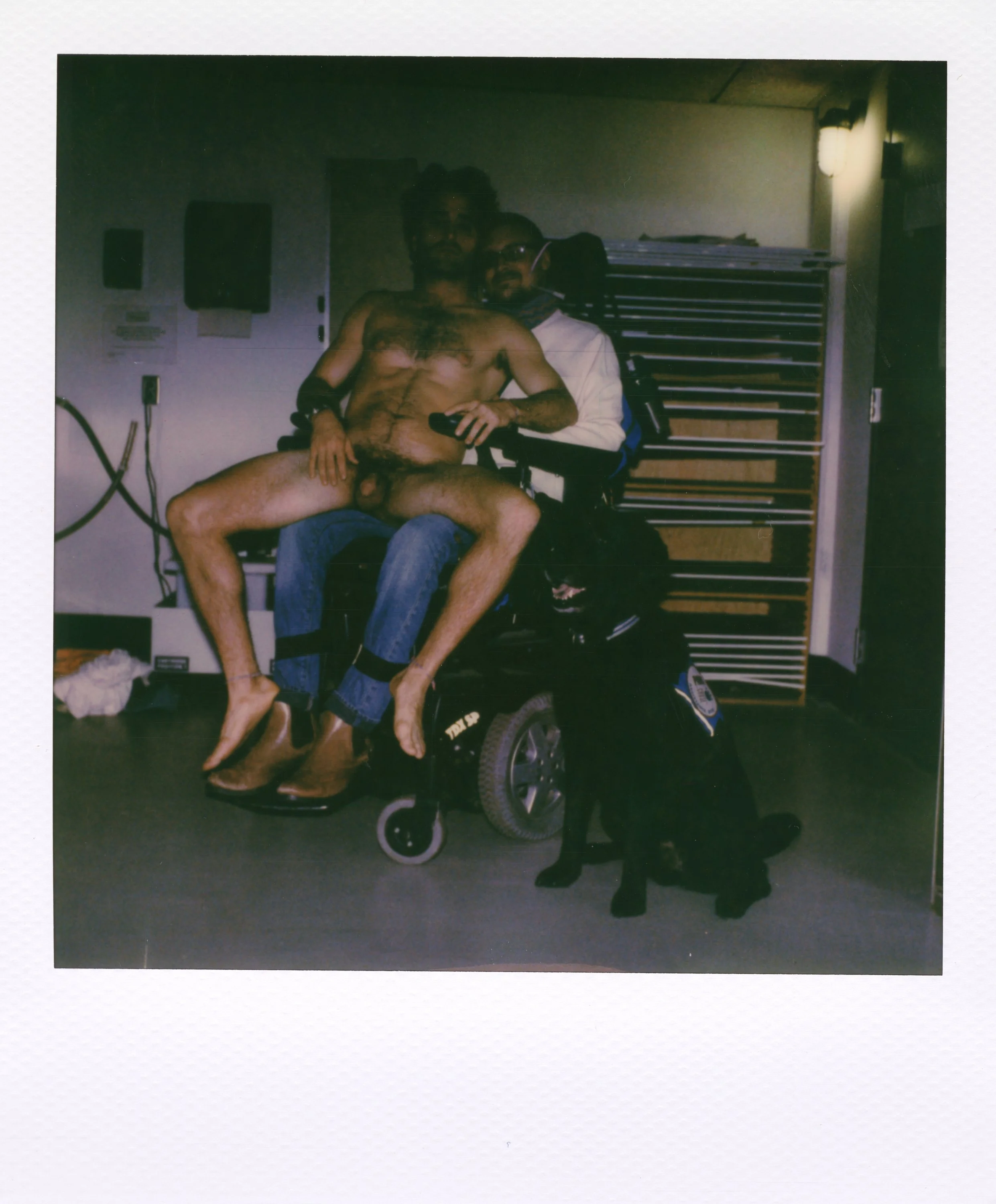 A man in a wheelchair holding a shirtless man with glasses and a beard on his lap, accompanied by a black guide dog wearing a harness, inside a room with a wooden-rack and wall sconce lighting.