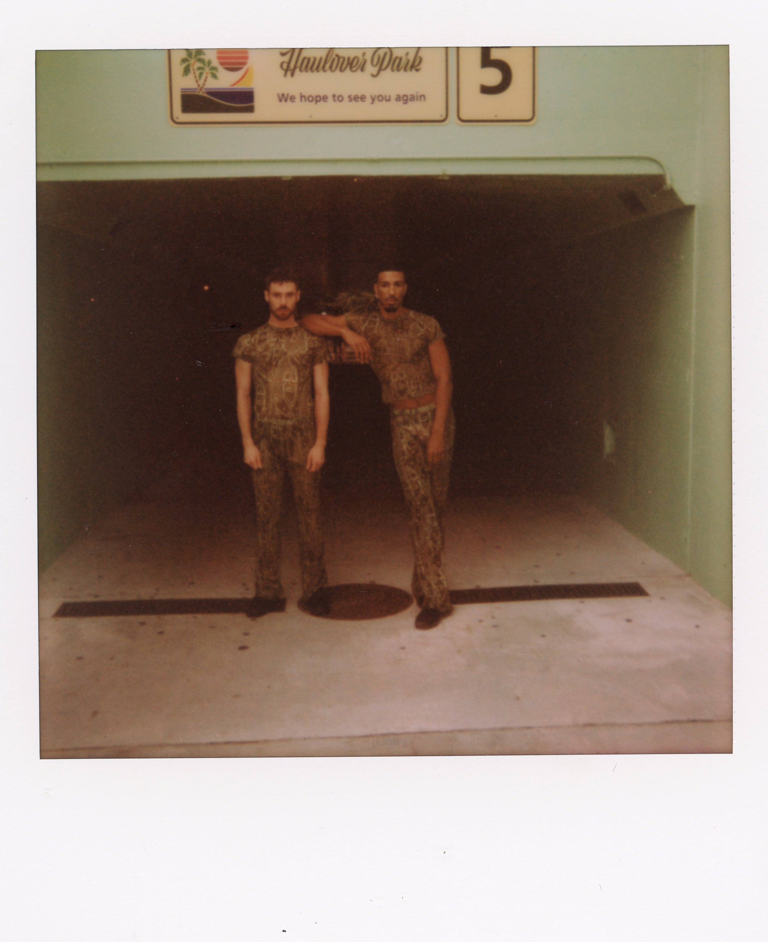 Two men in leopard print outfits standing beneath a parking garage entrance at night, with a sign above that reads 'Halloweer Park' and a number 5.