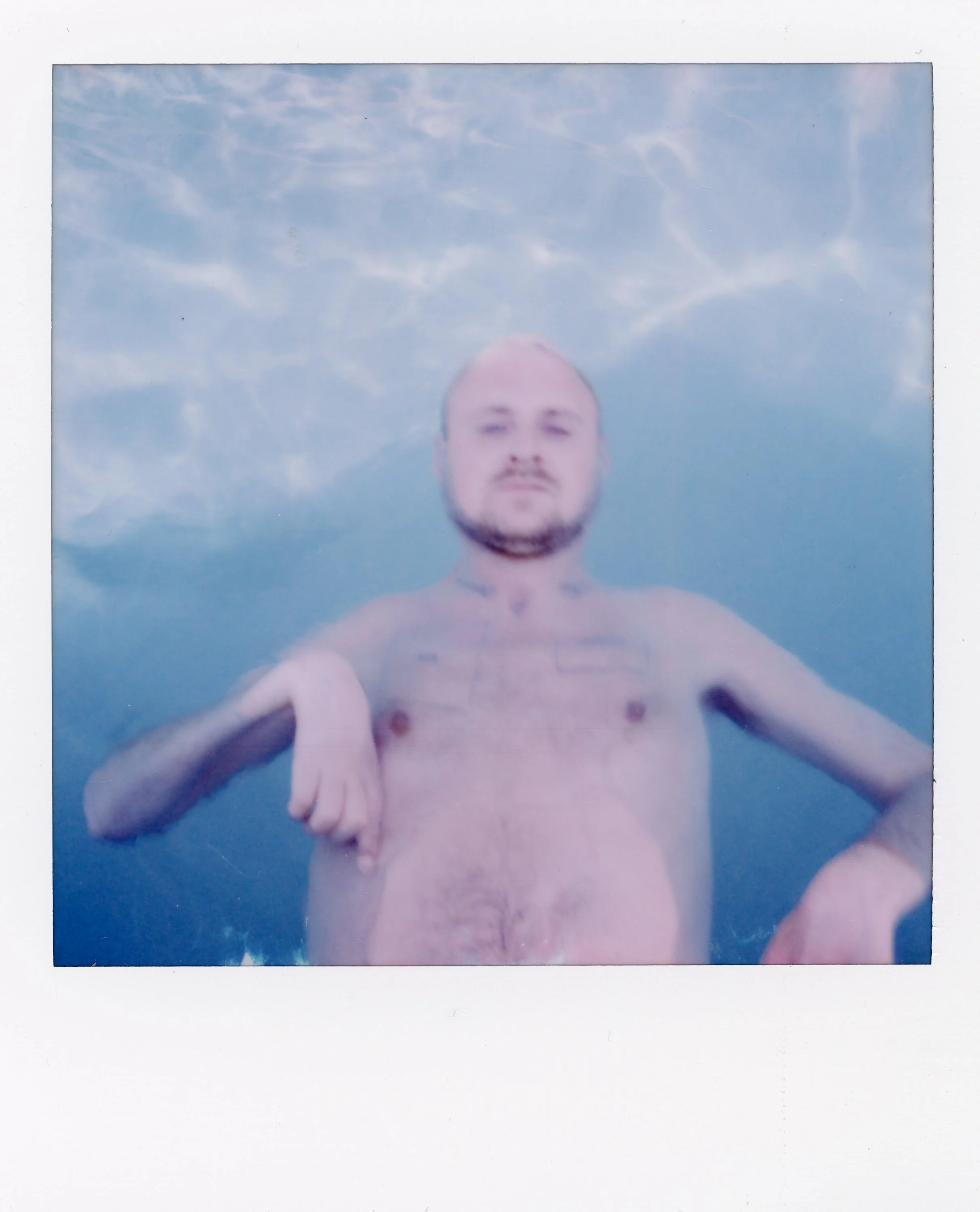 A shirtless man with a beard and tattoos, floating underwater with a cloudy sky above, looking directly at the camera.