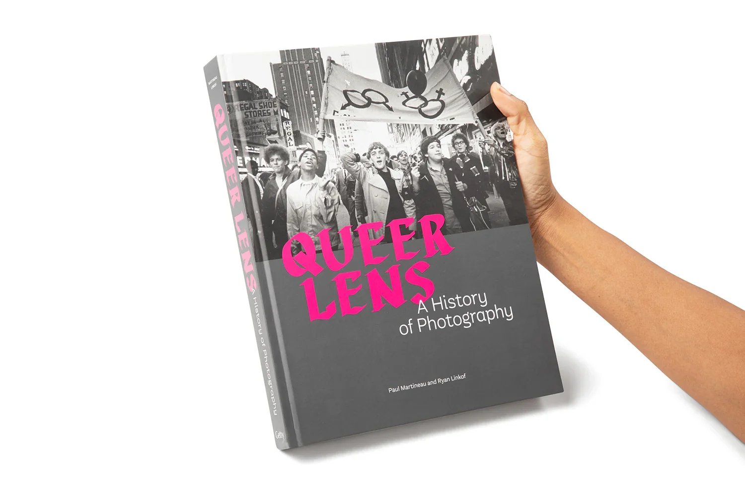 A person holding a book titled 'Queer Lens: A History of Photography.' The book cover features a black and white photograph of a parade or protest with diverse individuals, one holding a flag with gender symbols.