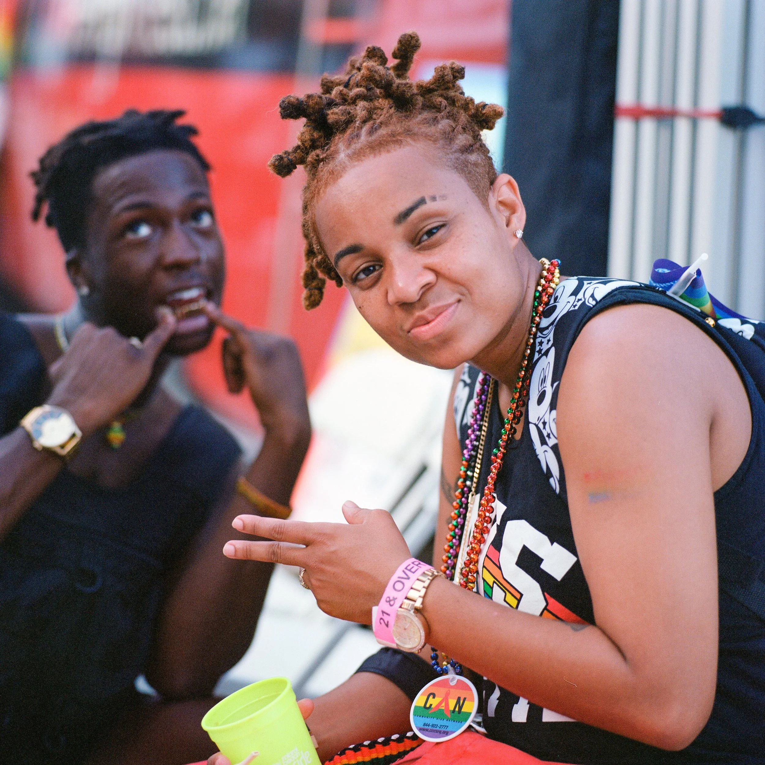 Two women at a pride event wearing colorful beads, smiling, with one making a peace sign and the other flashing a finger gesture, in front of an orange banner.