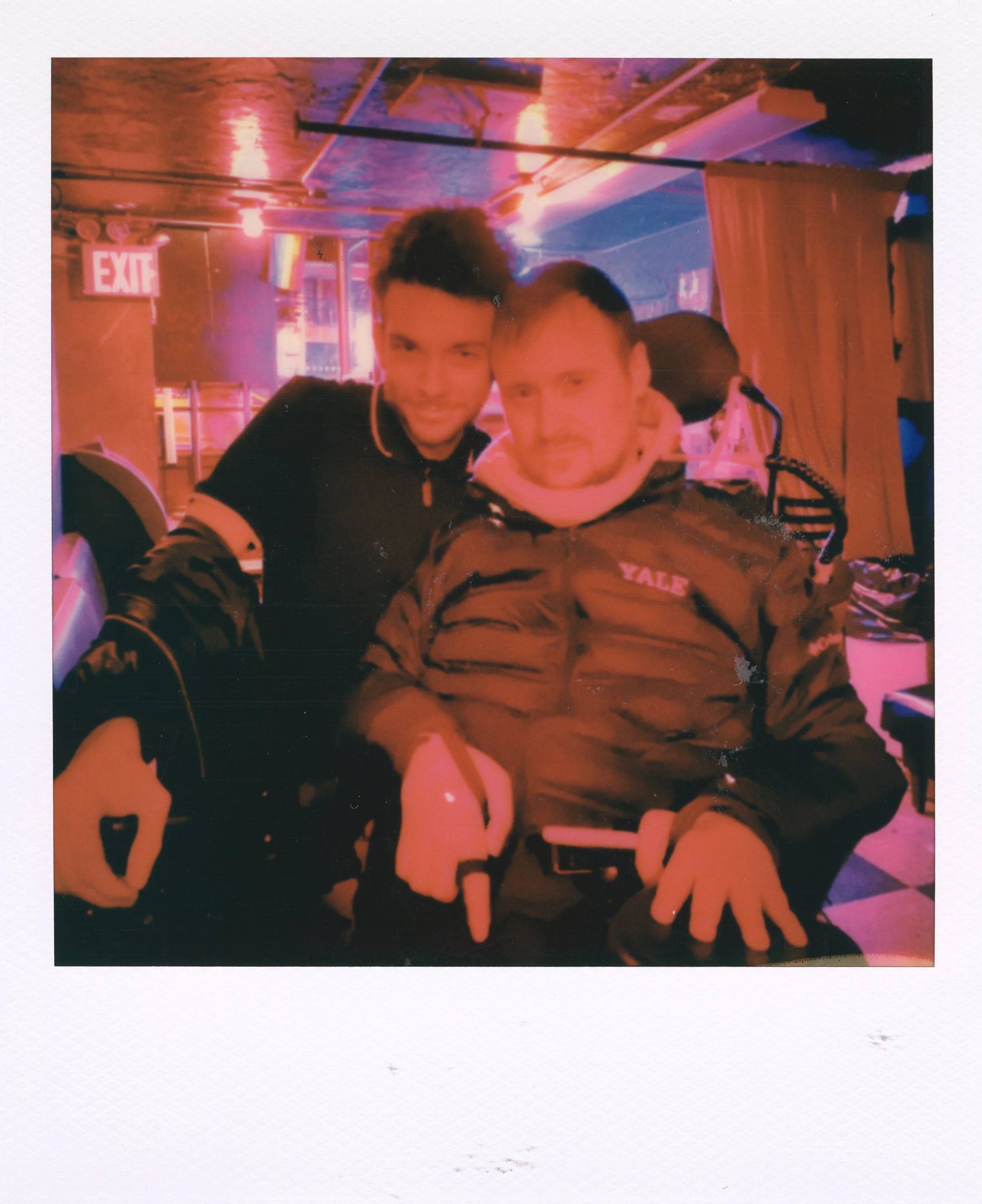 Two young men smiling, one in a wheelchair, in a dimly lit bar or club with pink and purple lighting, and an exit sign visible in the background.