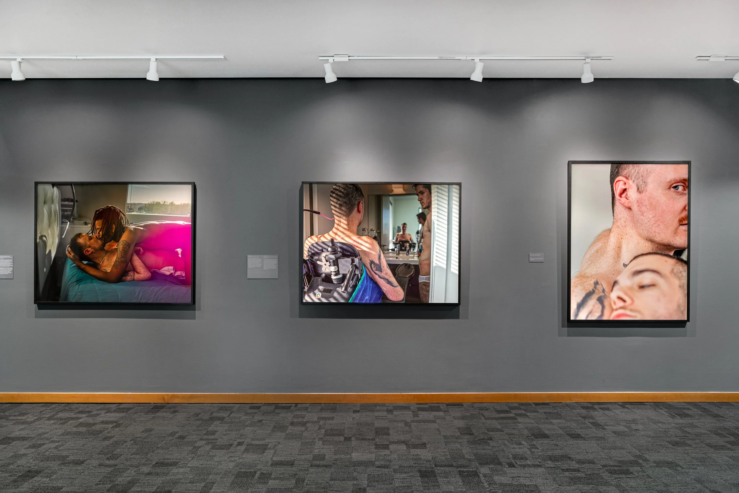 Three framed photographs on a gray gallery wall, depicting intimate candid moments of couples and individuals, featuring tattoos and close physical contact, with gallery lighting overhead.