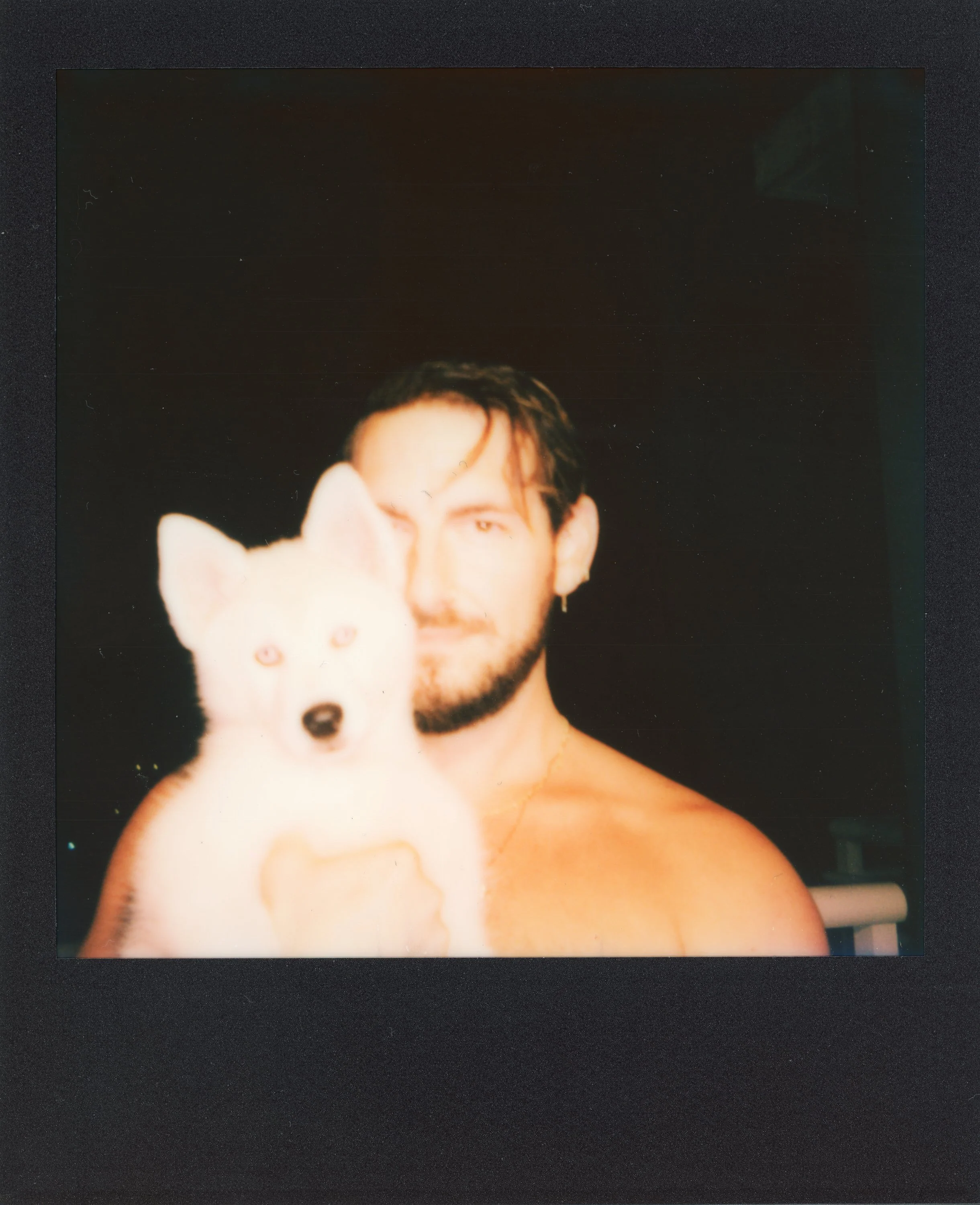 A shirtless man with dark hair and a beard holding a white dog with pointed ears, in front of a dark background.