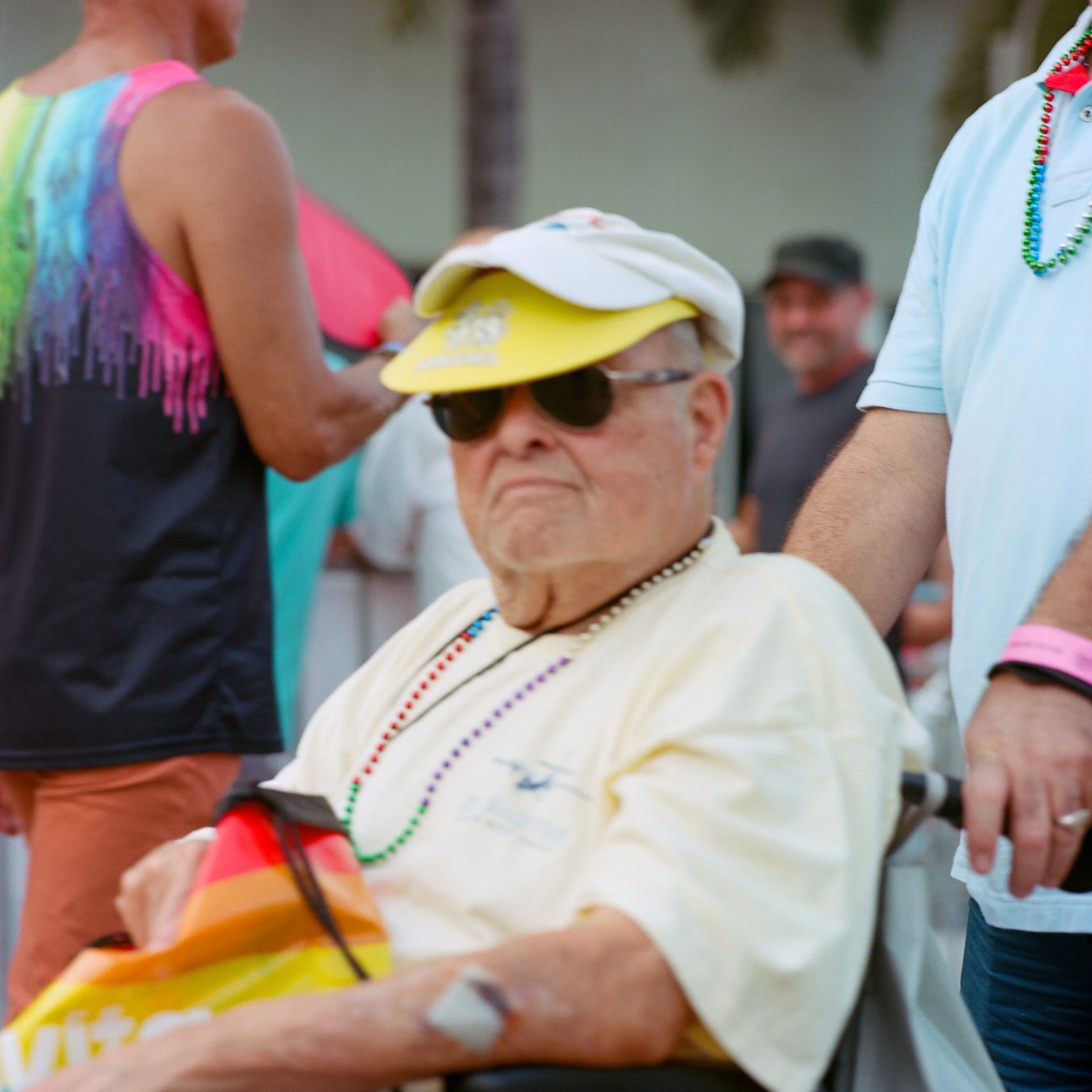 An elderly man wearing sunglasses, a beige shirt, colorful bead necklaces, a white cap with a yellow brim, and a white hat on top, sitting in a wheelchair at an outdoor event with other people around.