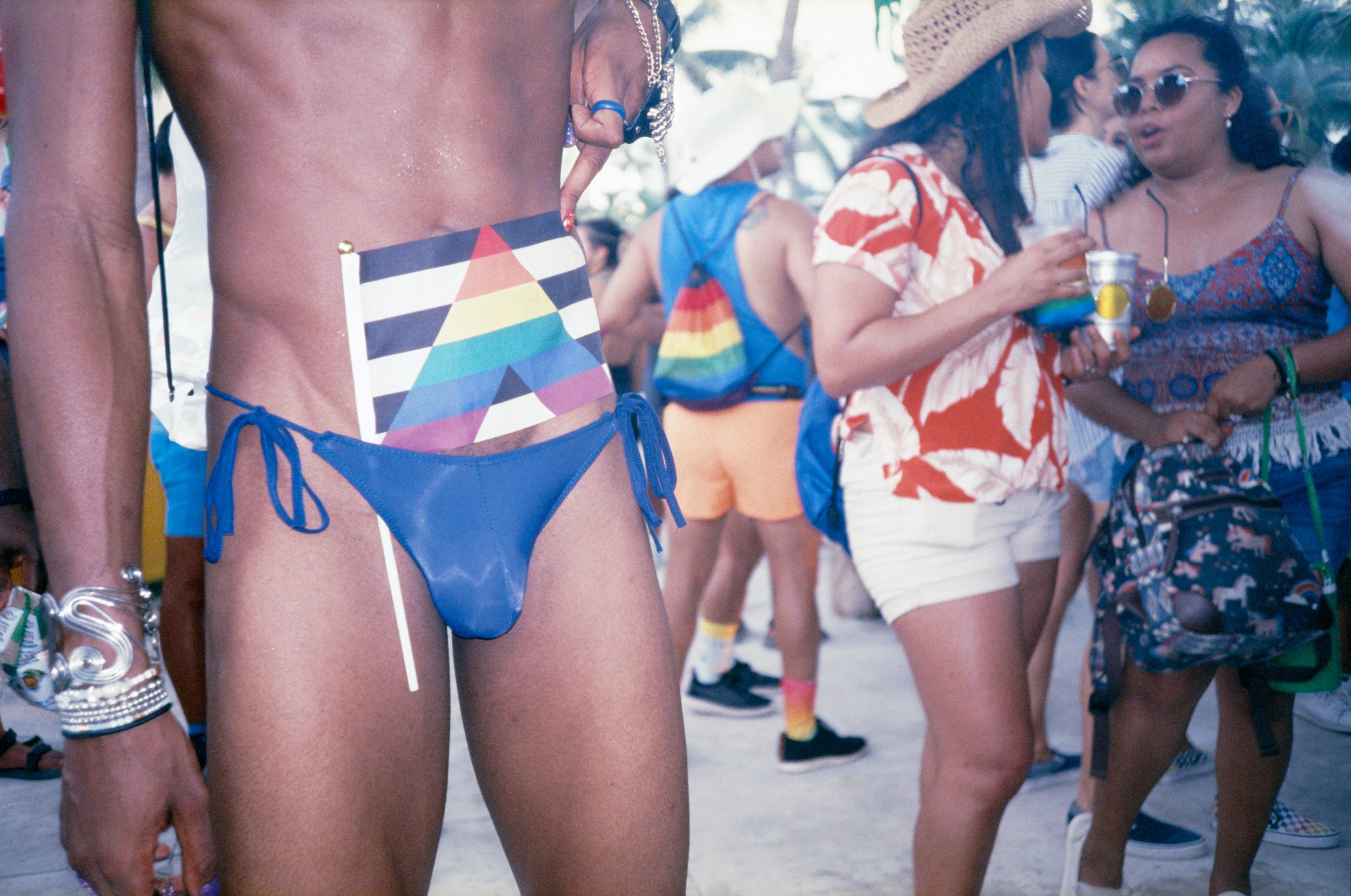 Close-up of a person in a blue swimwear with a rainbow flag tucked into the waistband, surrounded by people at an outdoor event, some holding drinks, with tropical plants in the background.