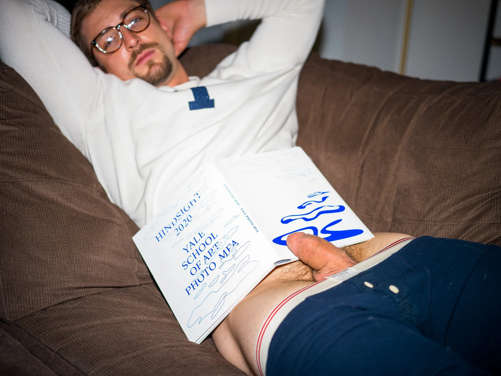 A man with glasses sleeping on a brown couch holding a Yale school of art 2020 photograph showcase booklet over his lap.