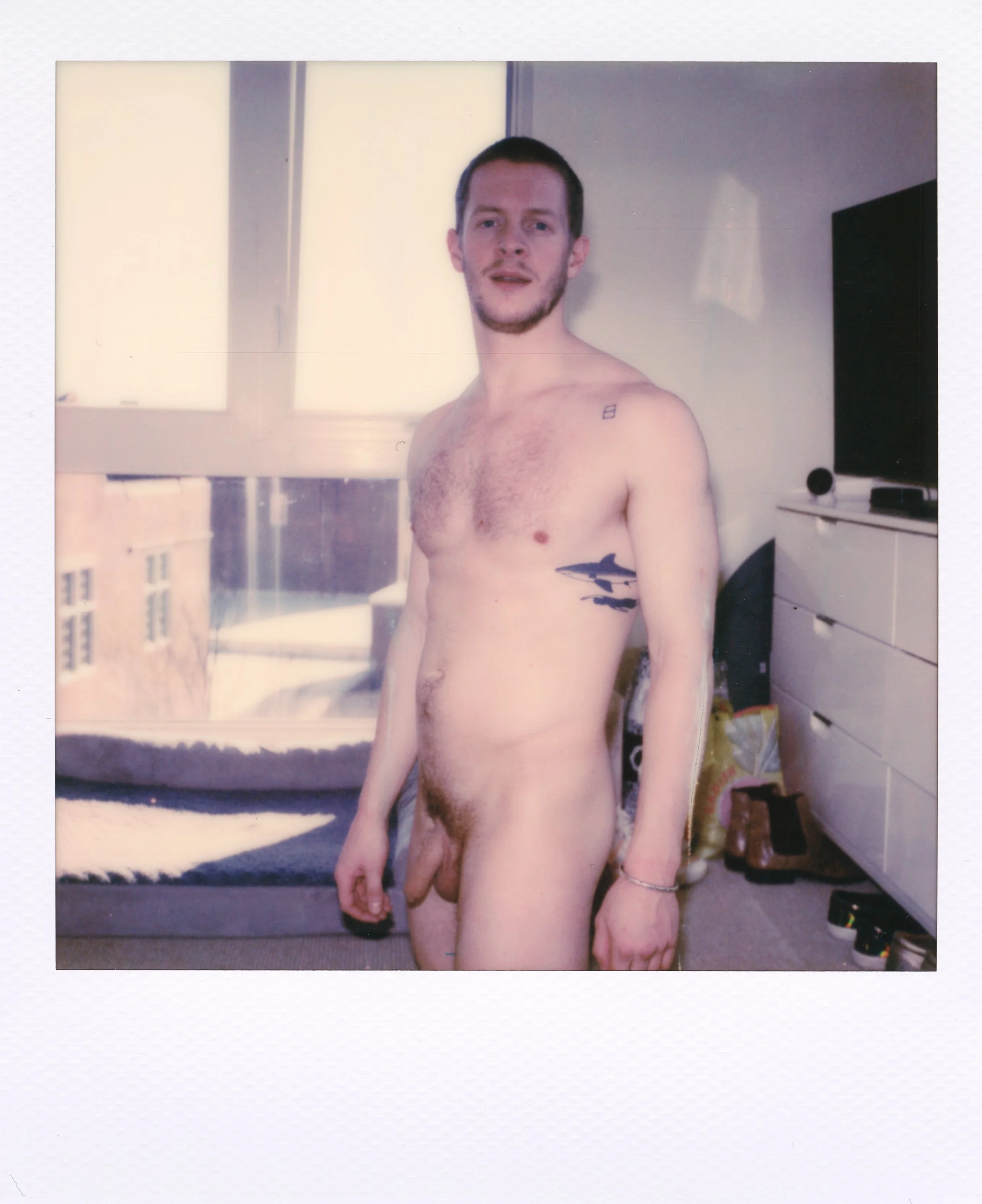 A young man with short dark hair, some facial hair, and tattoos on his left arm and side is standing in a brightly lit room, nude, looking at the camera. There is a bed, a dresser, and a flat-screen TV in the background.