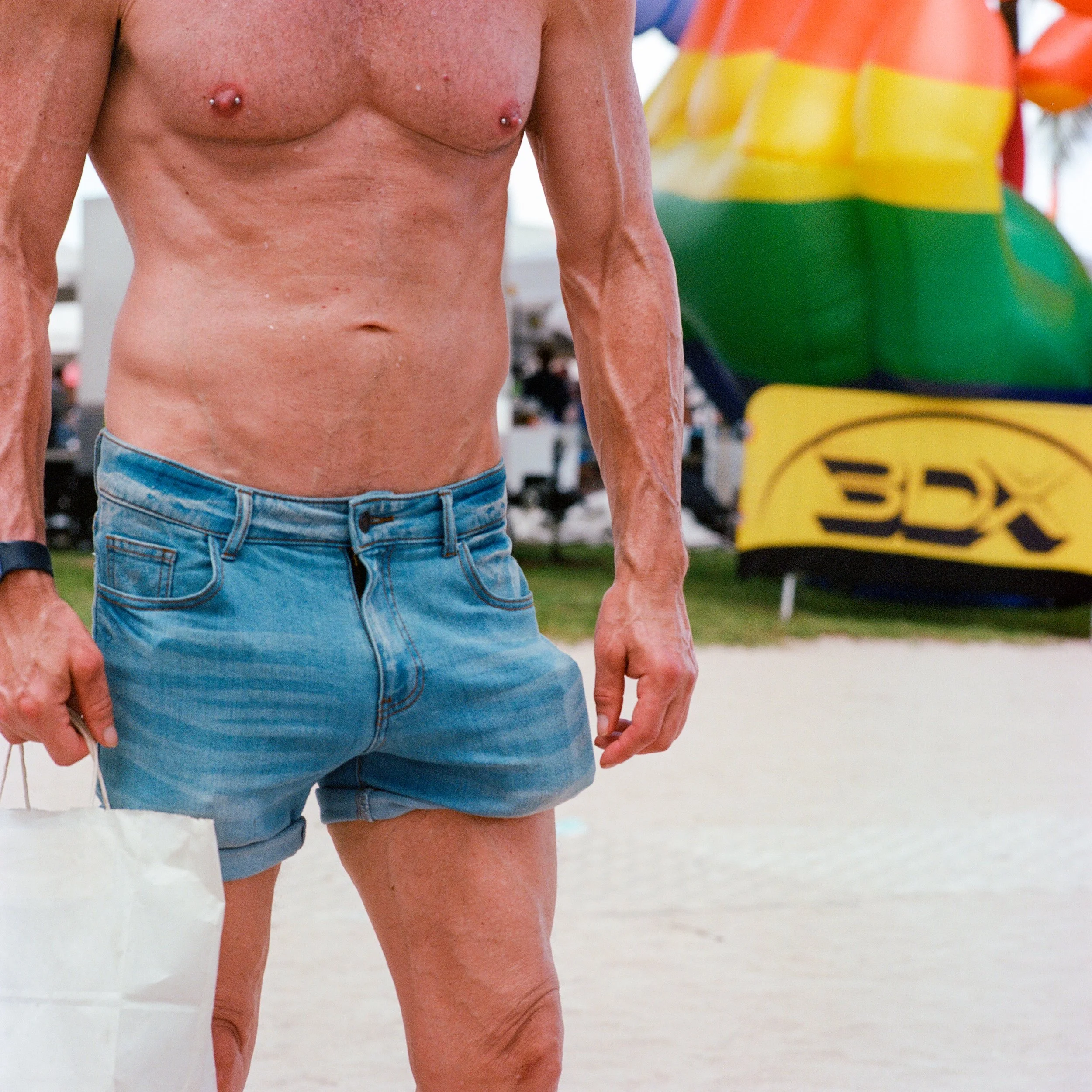 Close-up of a shirtless man wearing blue denim shorts holding a bag, with a colorful inflatable structure in the background.