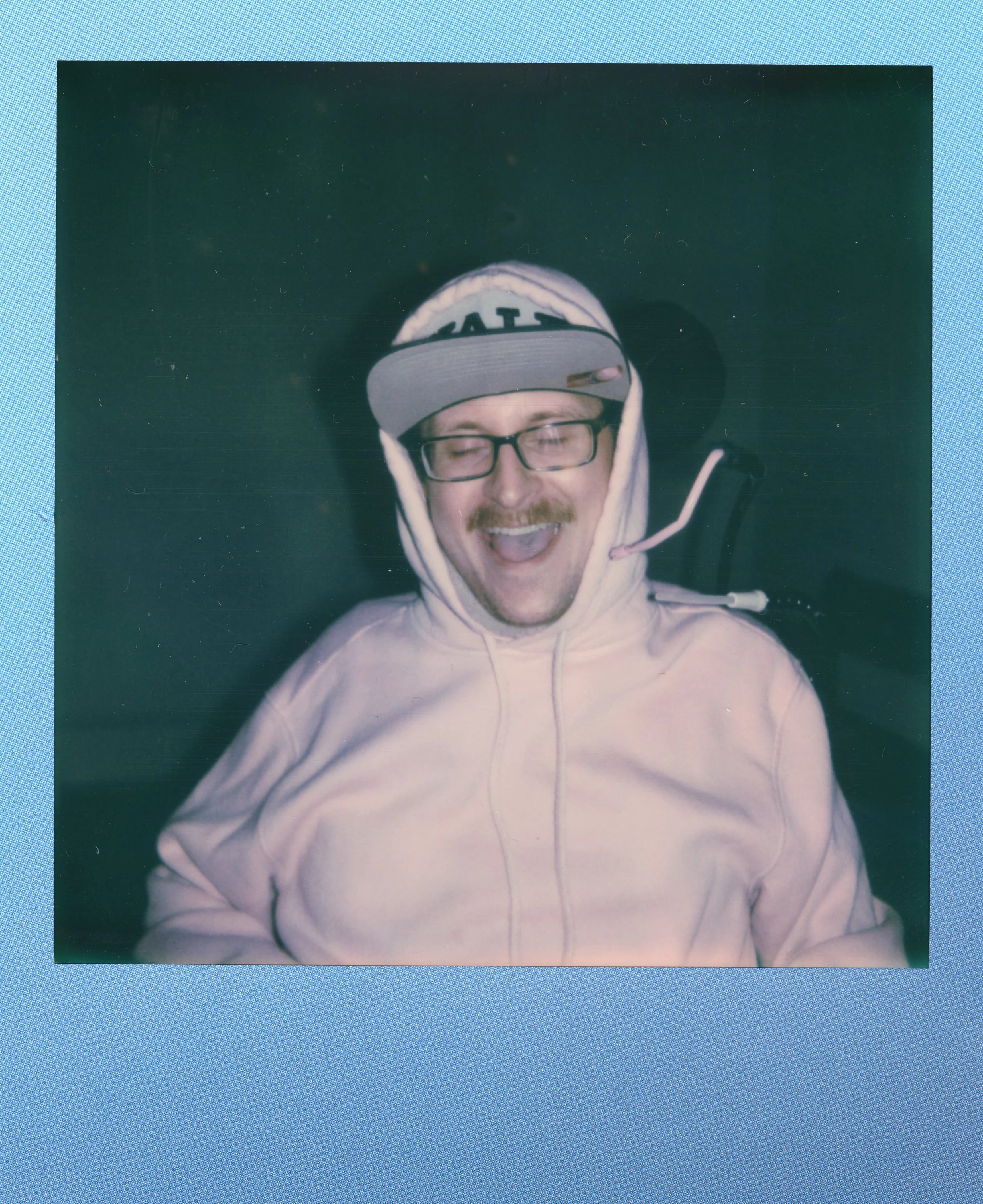 A man with glasses and a moustache wearing a white hoodie and a backwards baseball cap, with a headset on, smiling and laughing.