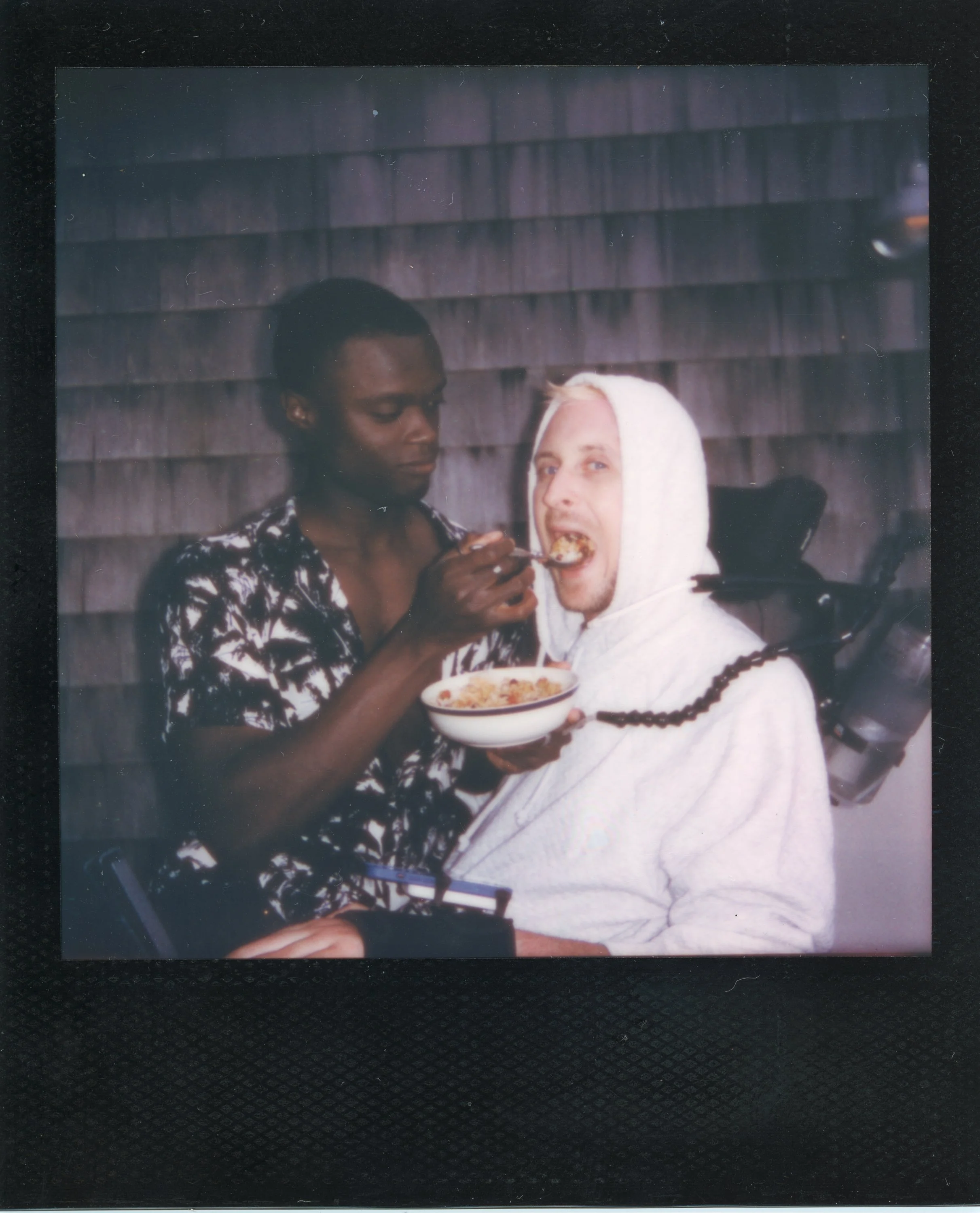 Two men, one with dark skin and the other with light skin, are sharing a bowl of food indoors. The man with dark skin is feeding the man with light skin, who is wearing a hoodie. The background shows wooden shingle siding.