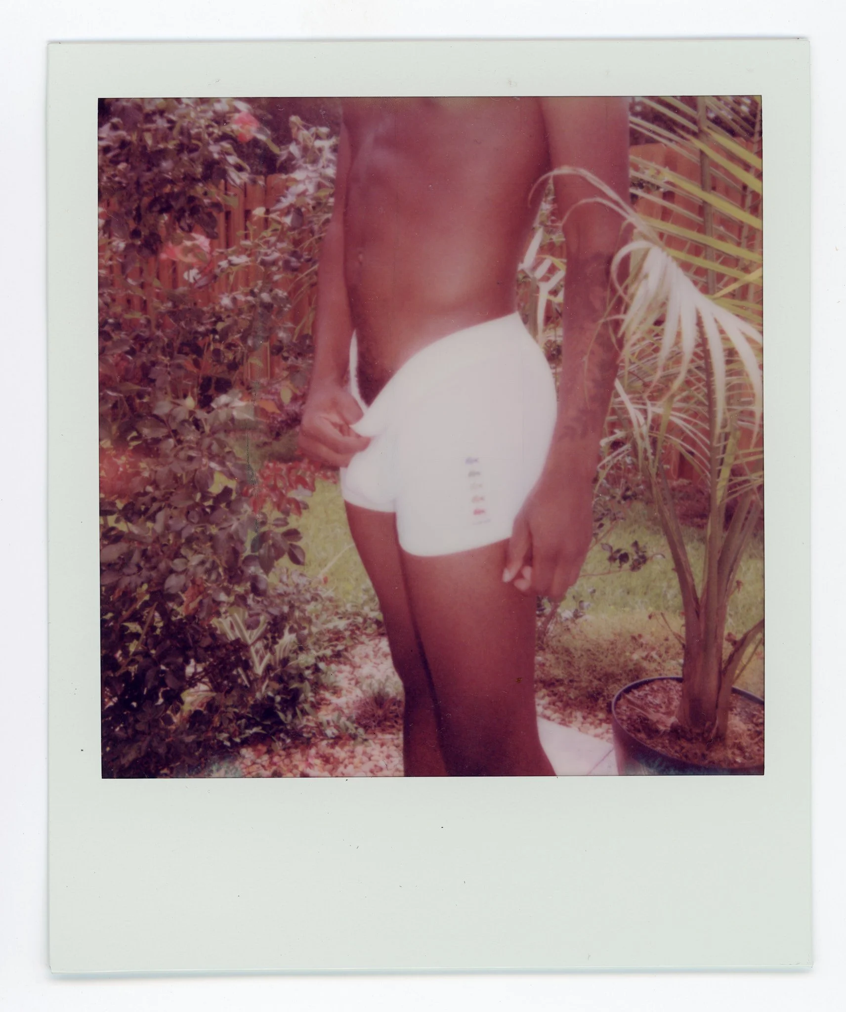A person with dark skin standing outdoors in a yard, only partly visible, holding a white handheld fan near their lower back, with plants and a wooden fence in the background.