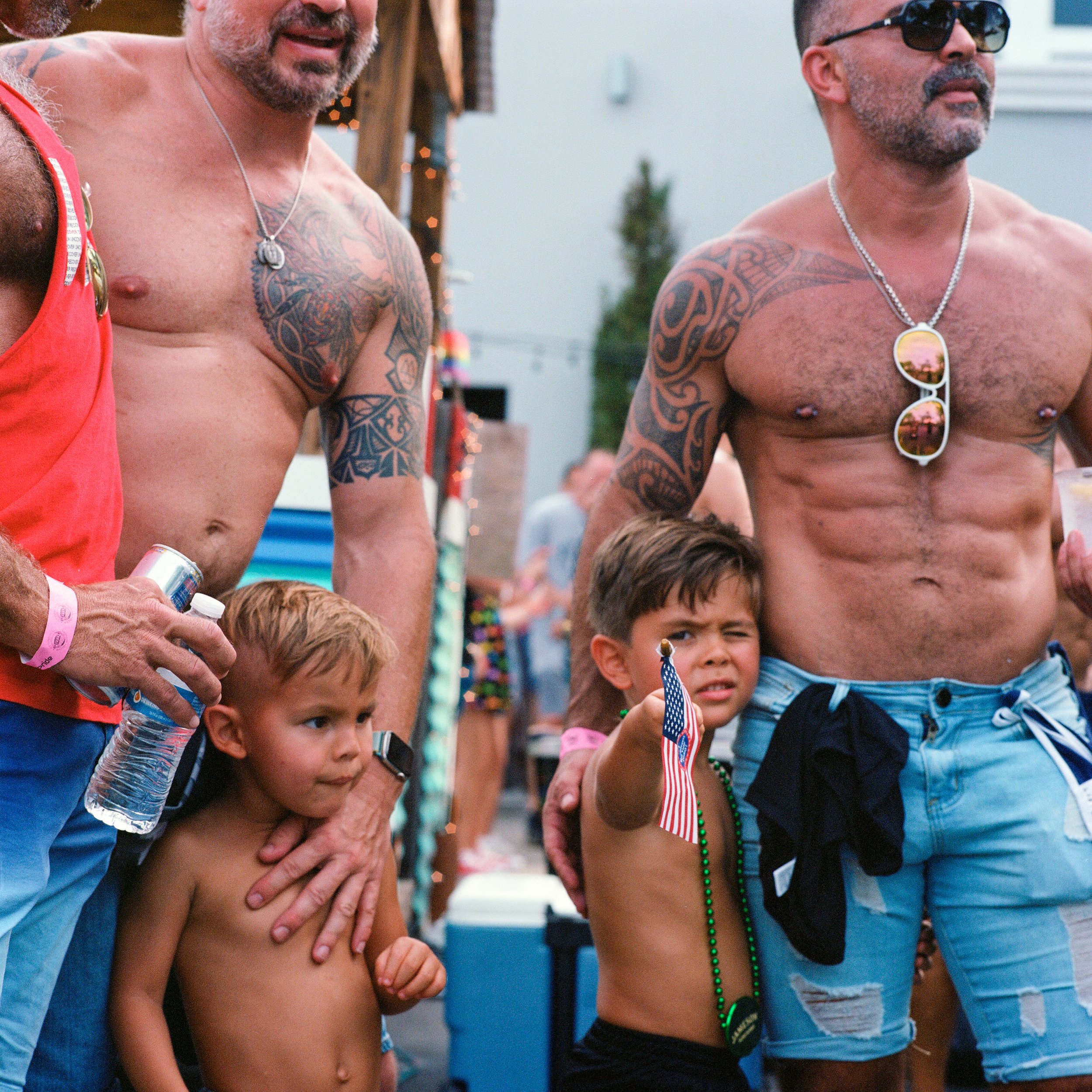 Two shirtless men with tattoos and sunglasses at a backyard gathering, each accompanied by a young boy; one man holding a water bottle, the other wearing sunglasses and holding a drink, with the boys standing close, one pointing with an American flag