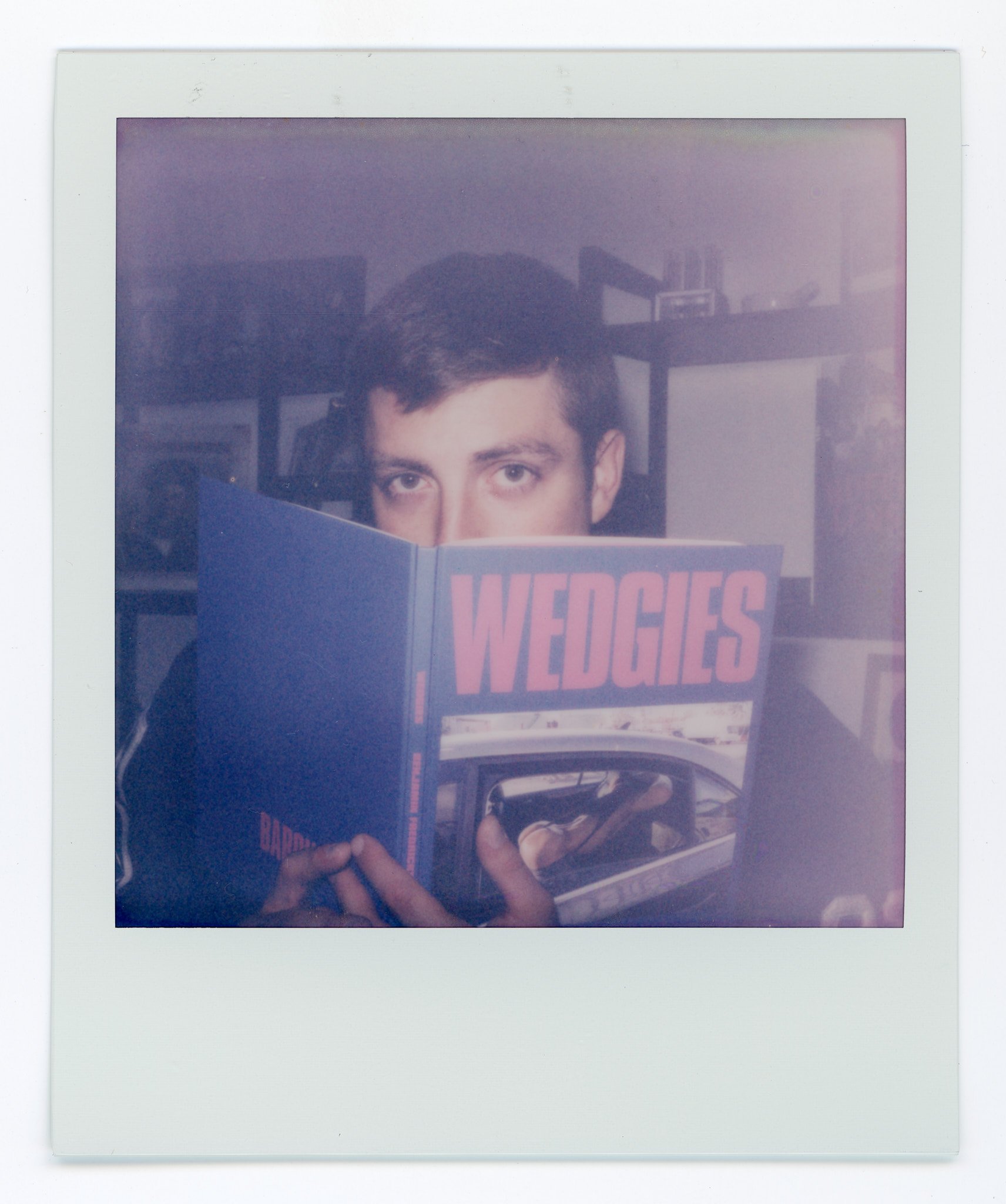A person reading a magazine titled "WEDGIES," with only their eyes and hands visible, in a room with bookshelf in the background.