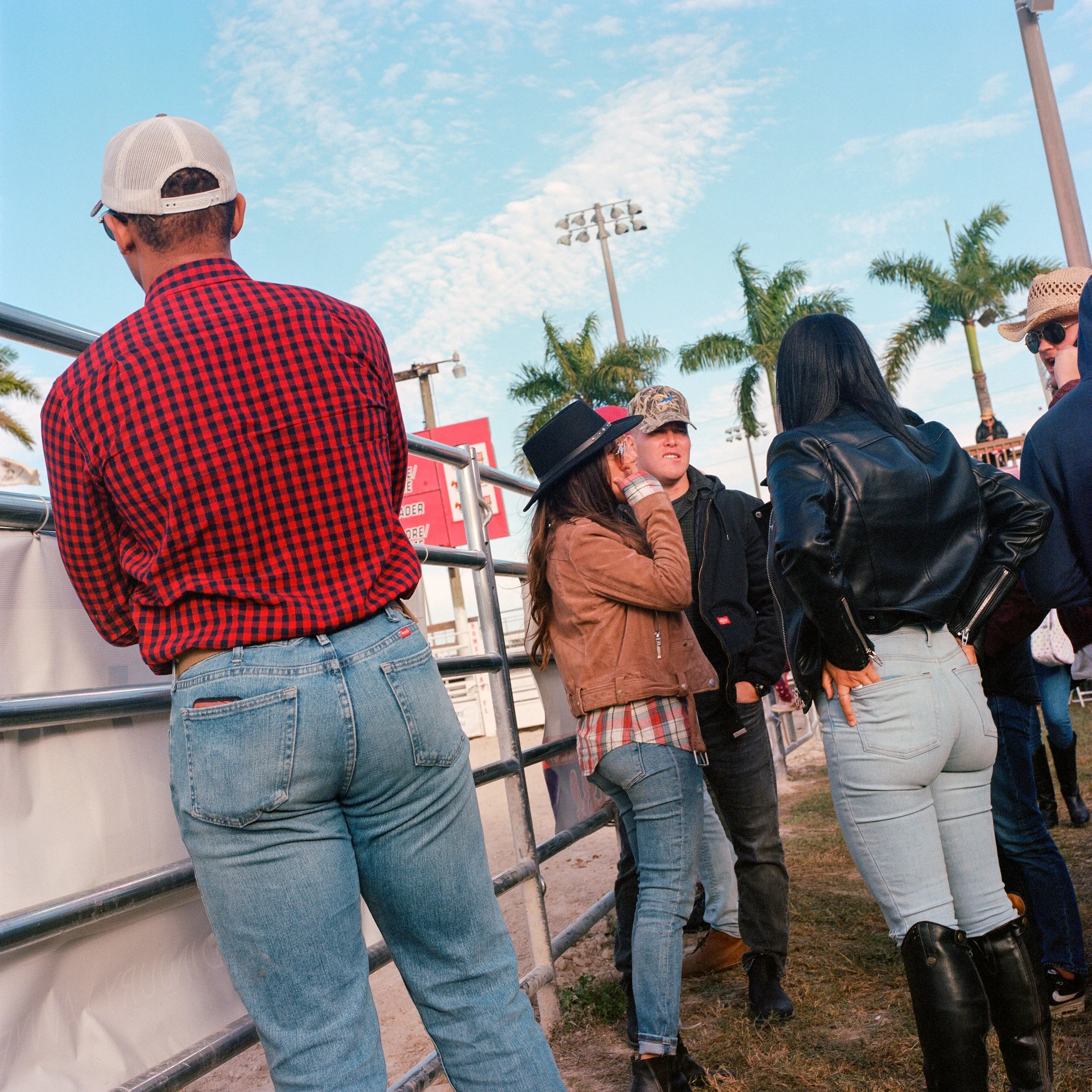 A group of people at an outdoor event, with palm trees and a blue sky in the background, some of whom are wearing hats and jackets, standing near a metal barricade.