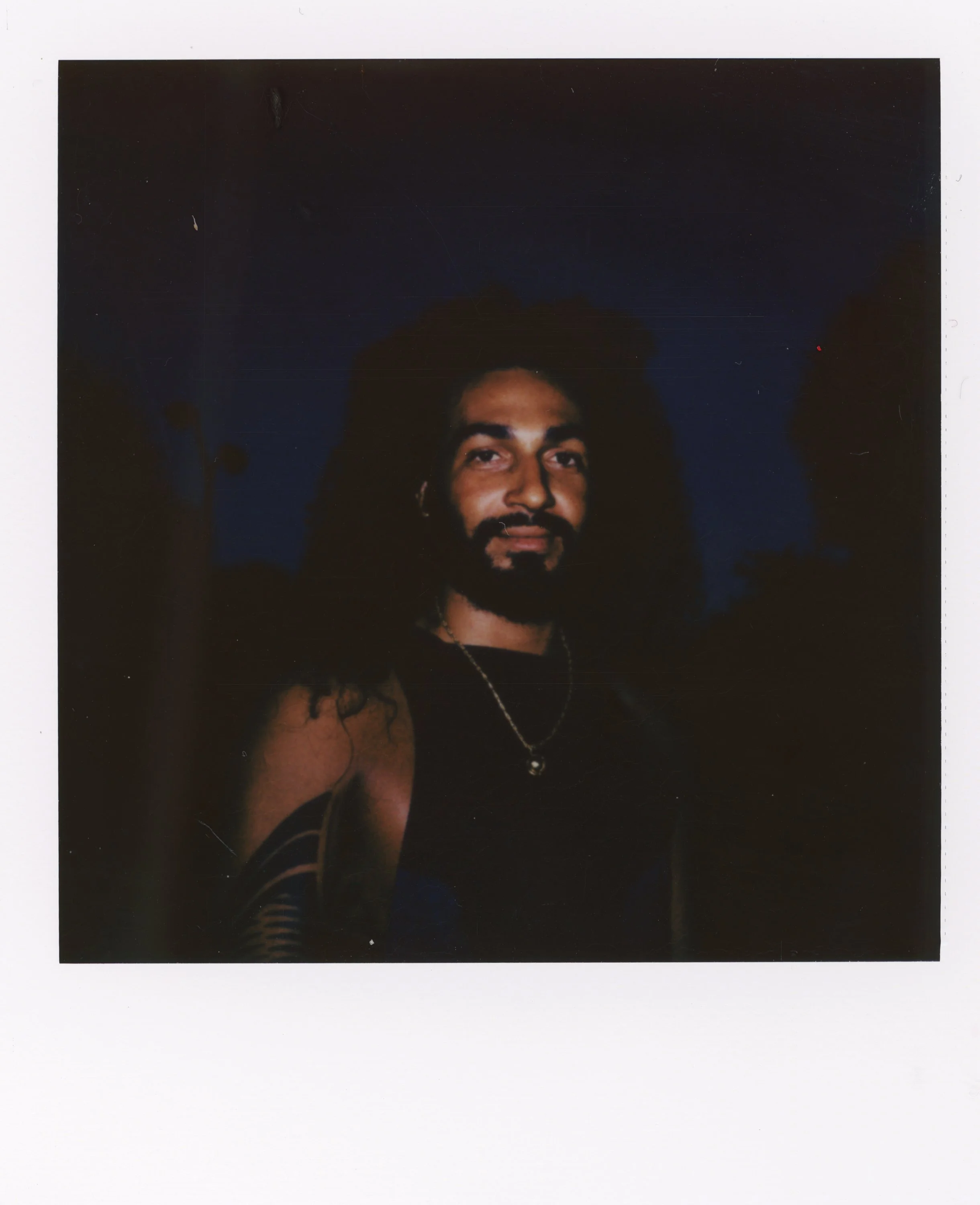 A man with long curly hair, a beard, and mustache, wearing a black sleeveless top, a necklace, and sitting outdoors at dusk or night.