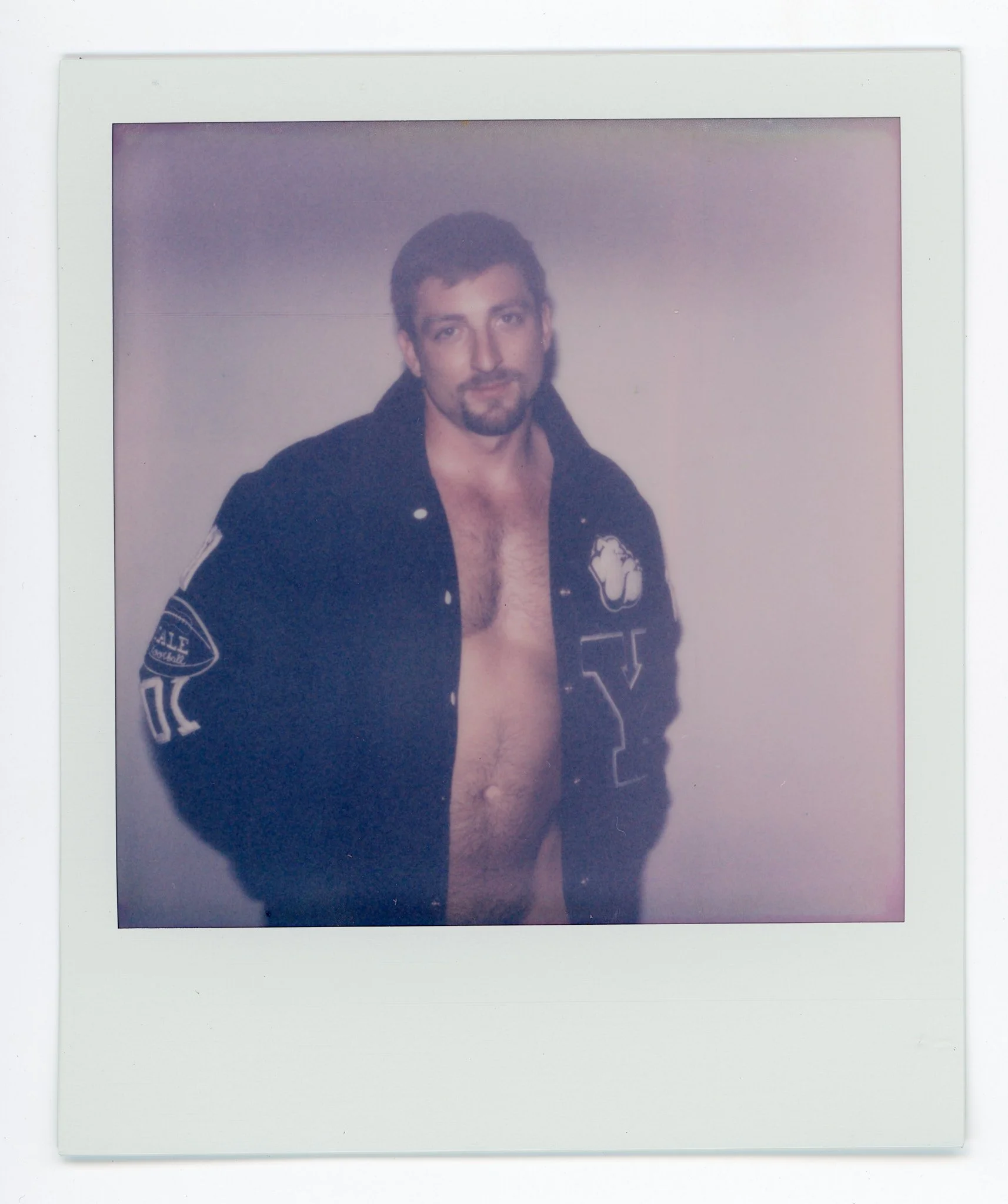 A shirtless man with a beard and mustache, wearing a black jacket with patches, standing against a plain wall.