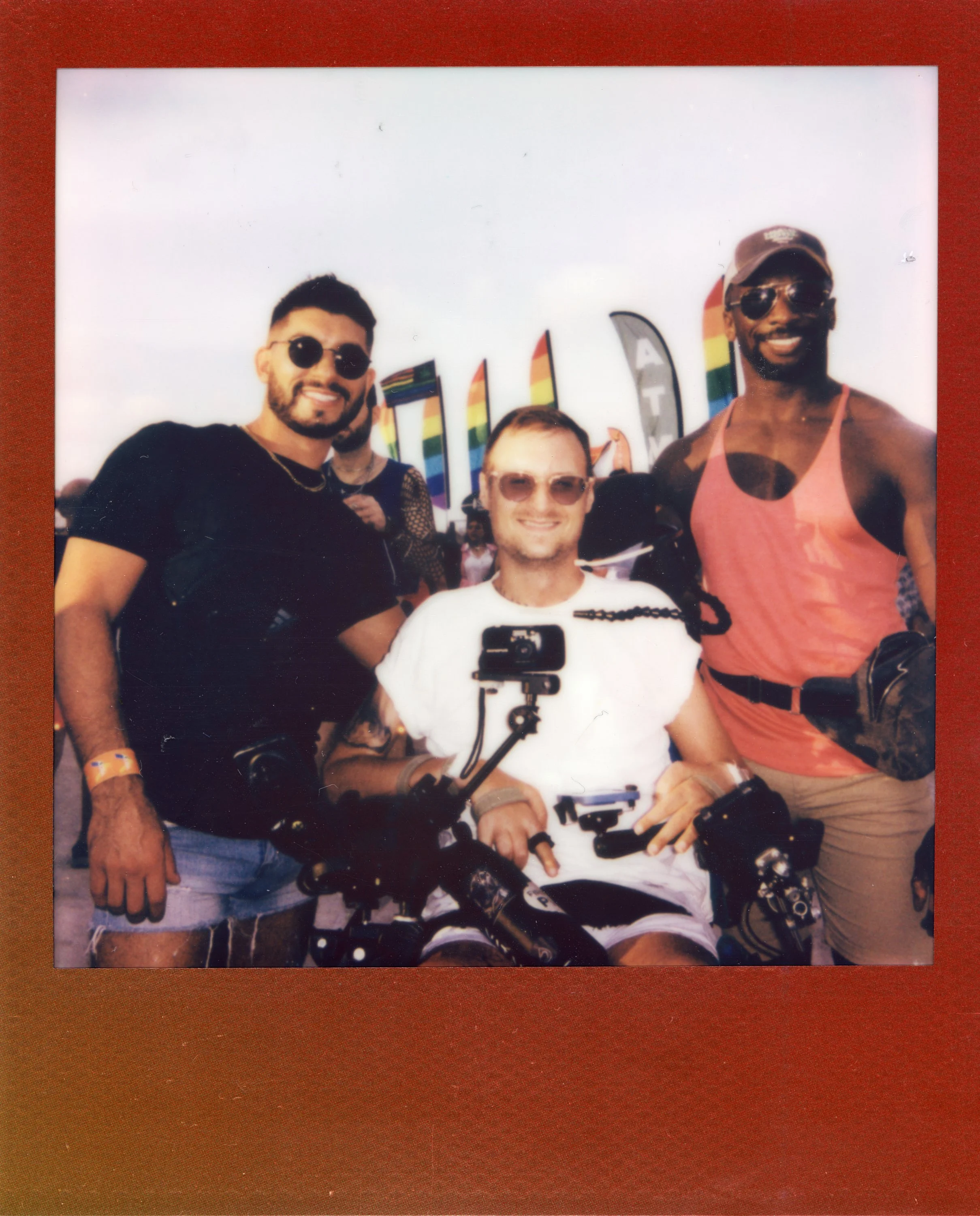 Three smiling men at a pride event, with rainbow flags in the background. The man in the center is in a wheelchair with equipment attached, and the other two men are standing beside him, all wearing casual summer clothing.
