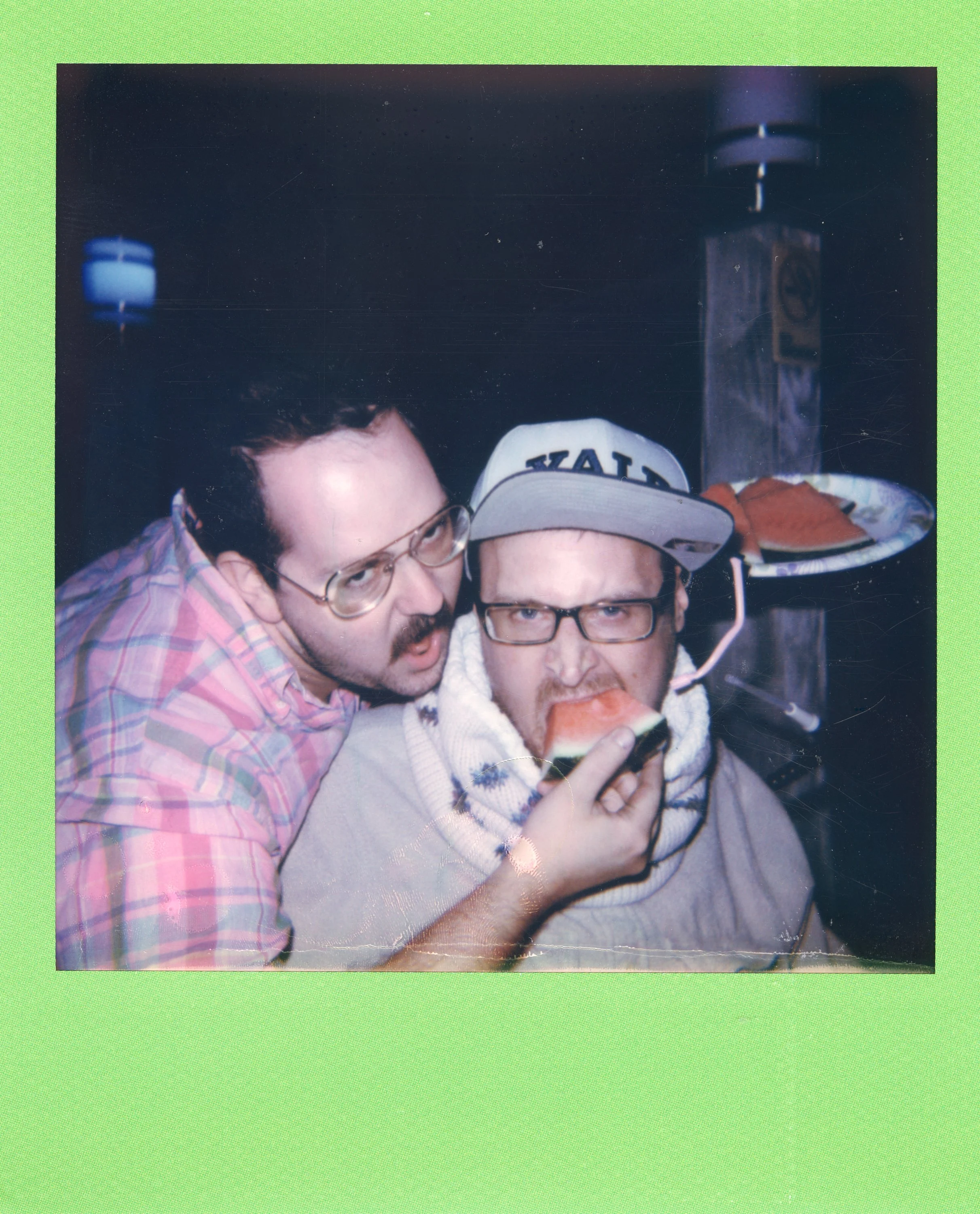 Two men with glasses sharing a watermelon slice with one man biting into it.