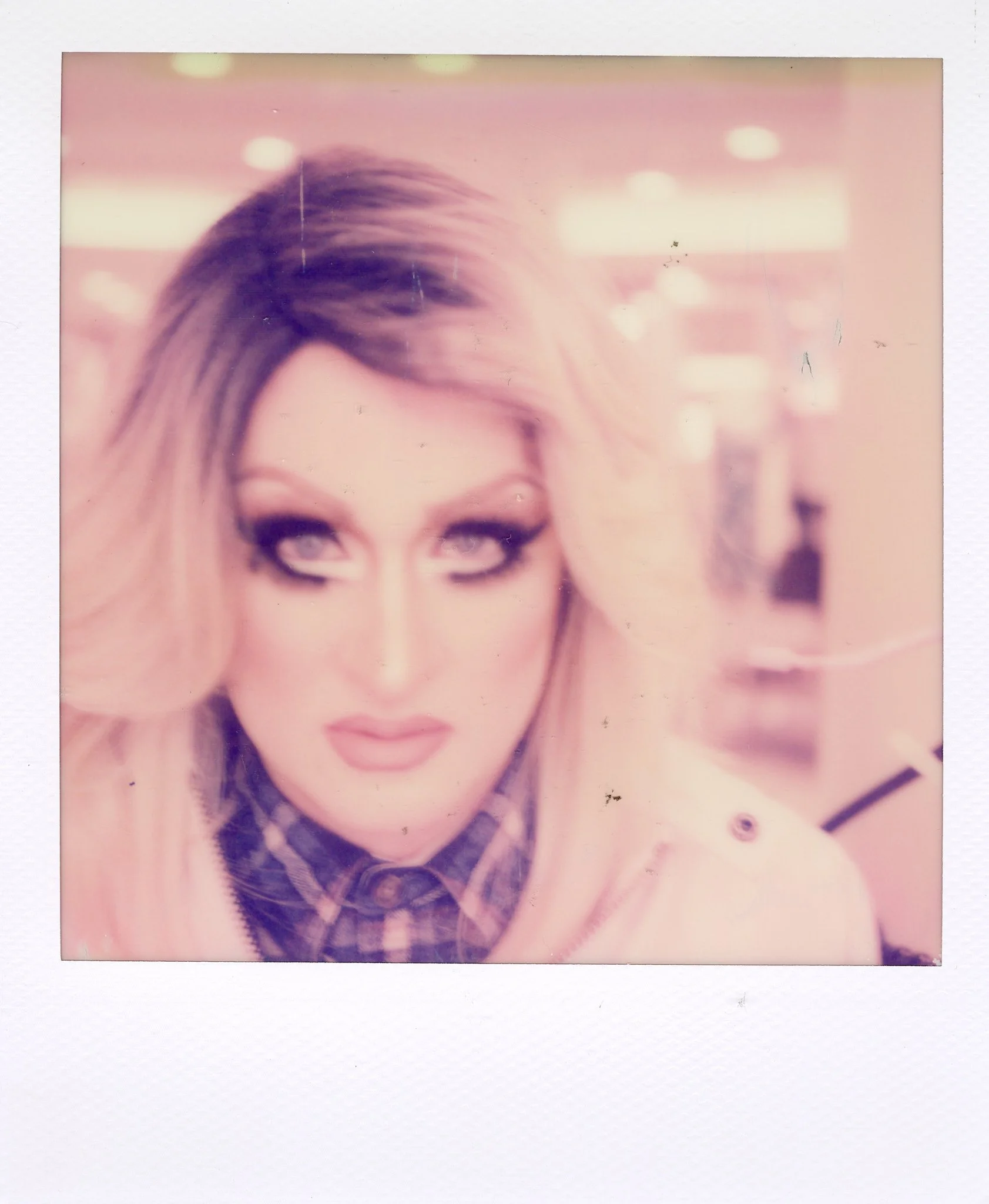 A woman with platinum blonde hair and makeup, wearing a plaid shirt and a light-colored jacket in a blurred, pink-tinted background.