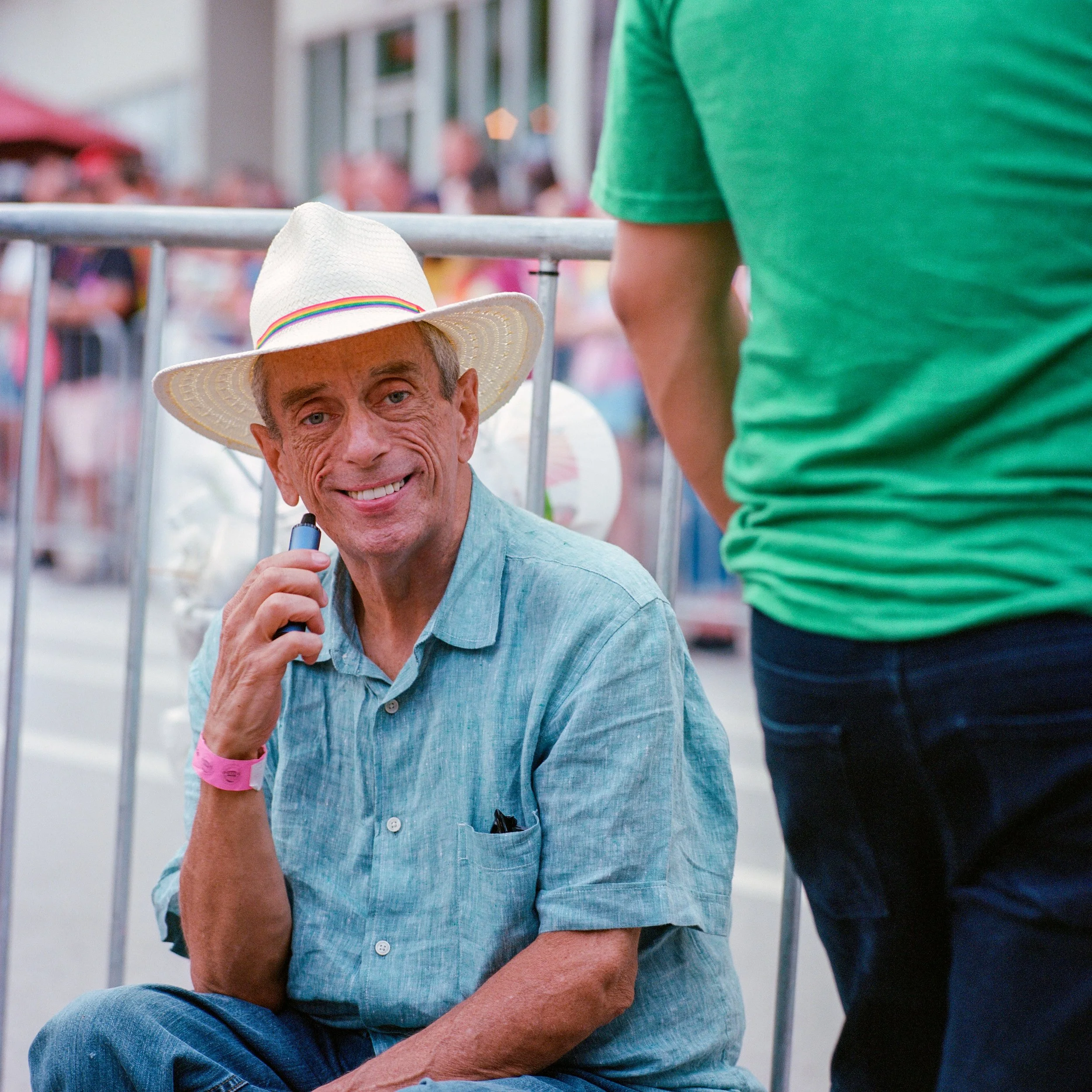 A man wearing a white hat with a rainbow band, blue shirt, and pink wristband smiling and holding a small electronic device, sitting outdoors near a metal barrier with a crowd in the background.
