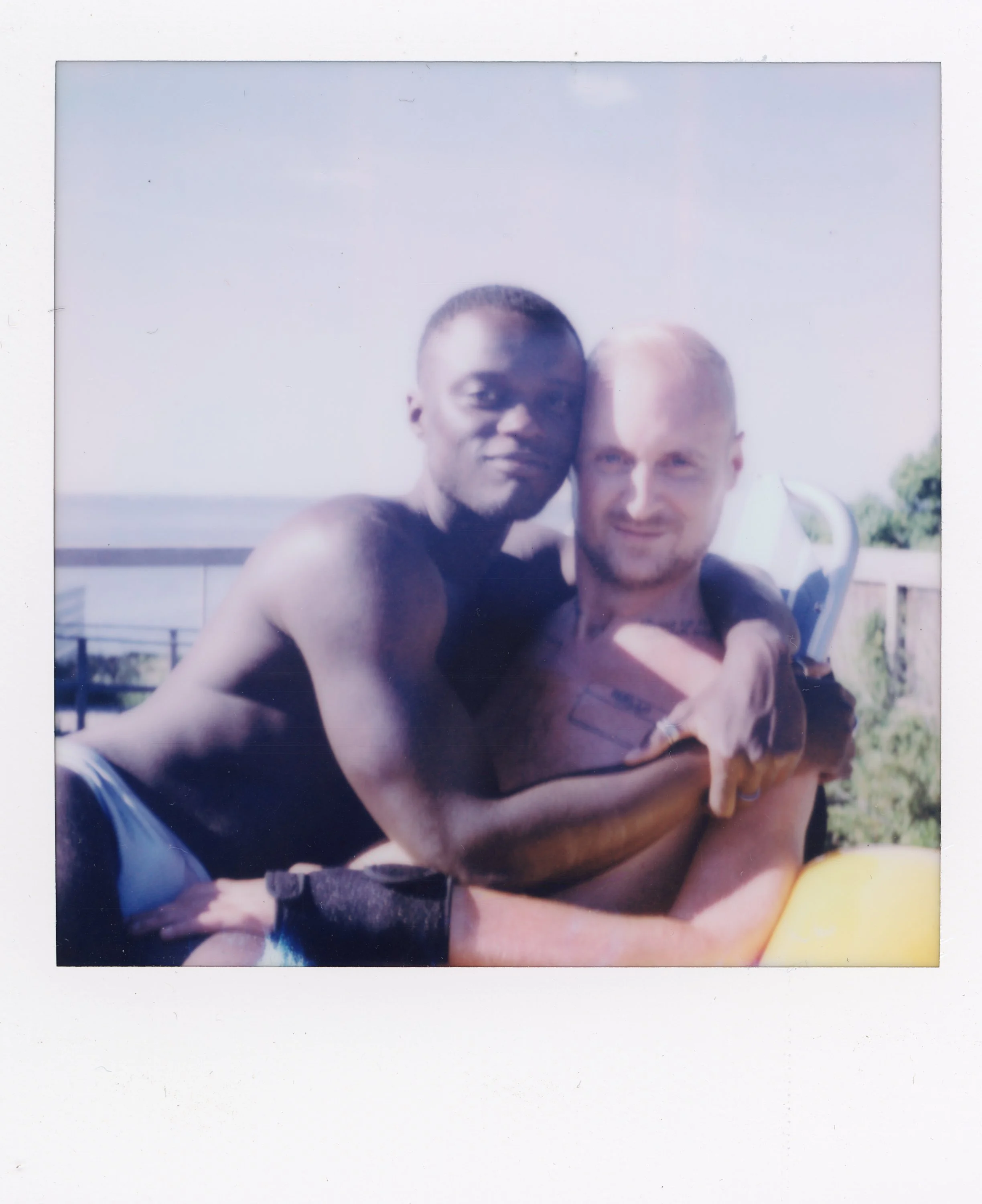 Two young men sitting outdoors, one with dark skin and short hair, the other with light skin and blonde hair, embracing each other and smiling at the camera, with a railing and trees in the background.