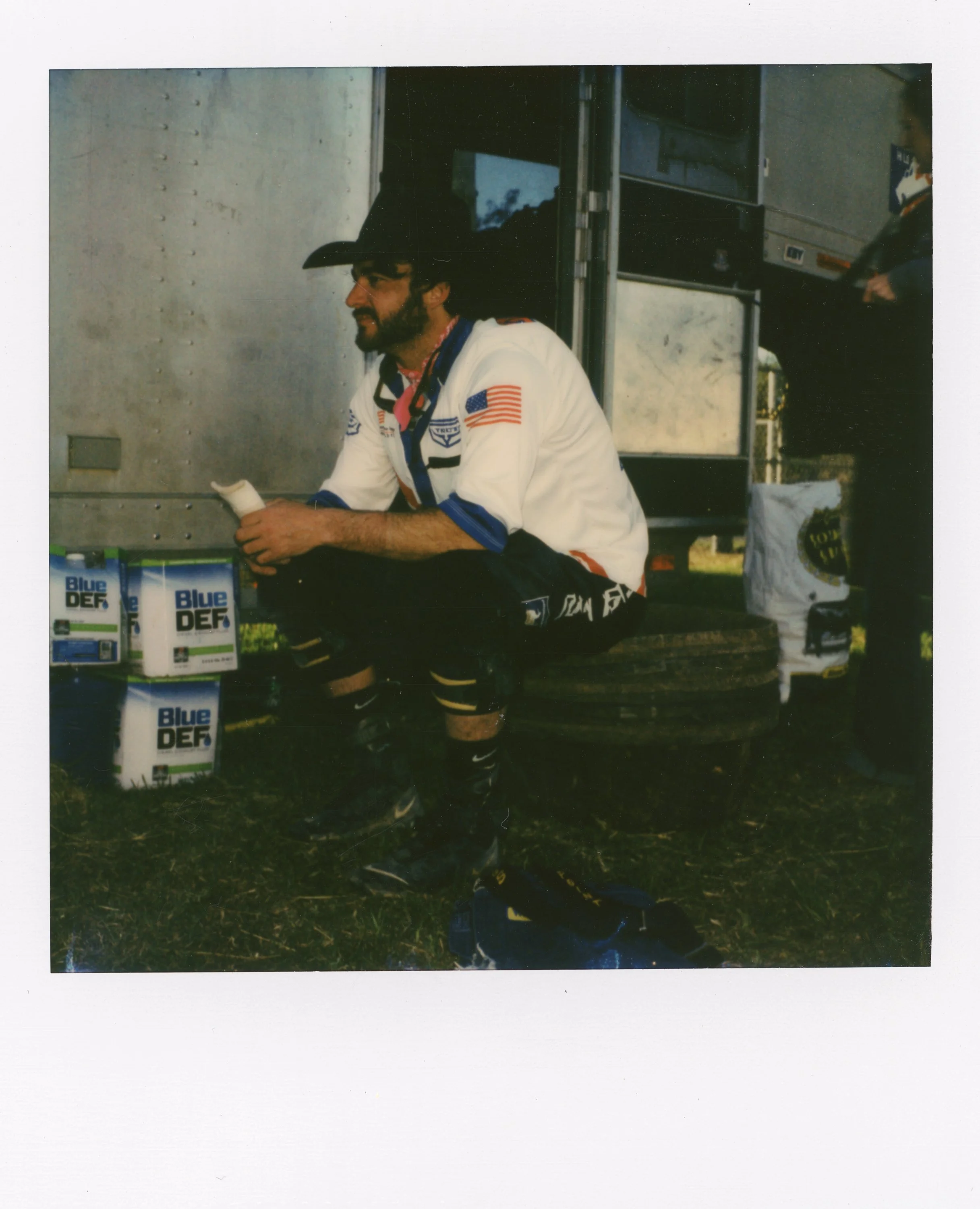 A man dressed in racing gear, sitting on a tire, holding a white object, with a trading card nearby, outside near a trailer.